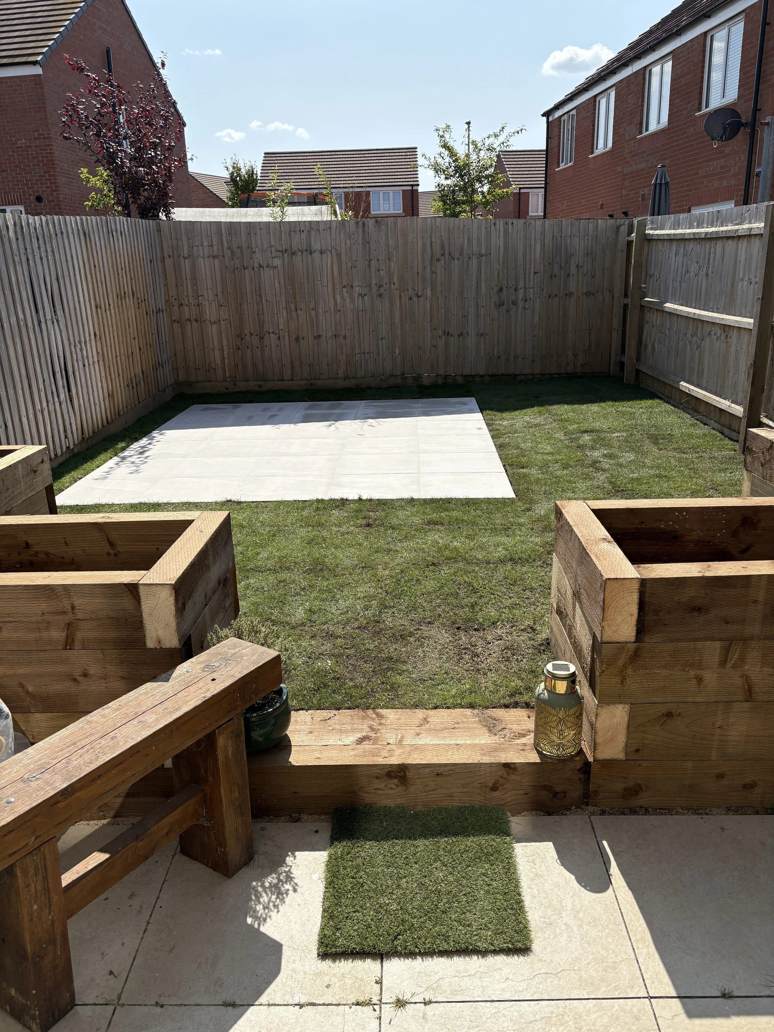Backyard with a wooden fence, a small patch of grass, a white tiled area, and a wooden planter box in the foreground. Sunny weather with a few clouds in the sky.