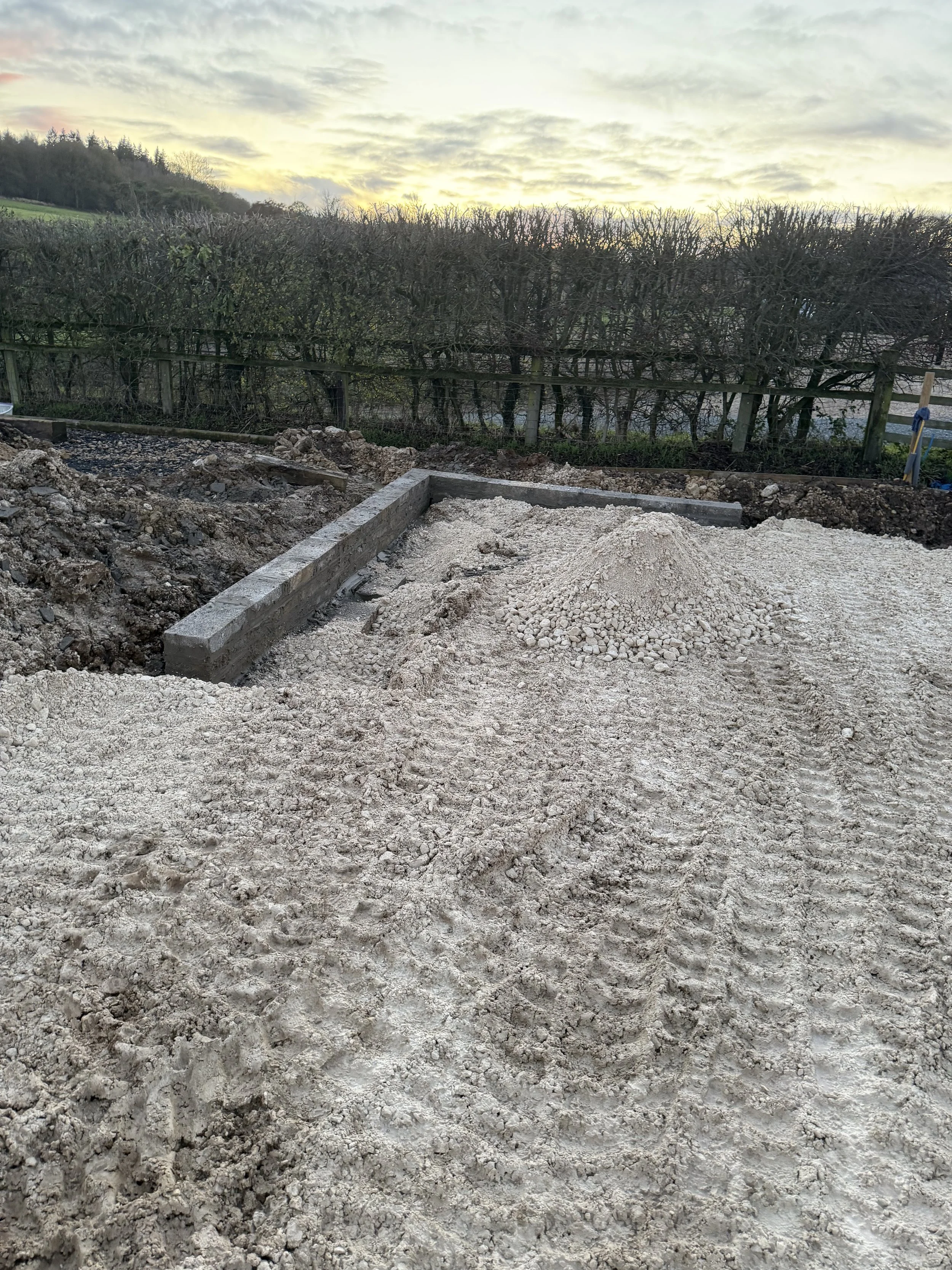 Construction site with a concrete foundation wall and piles of gravel and dirt, backed by a hedge and trees at sunset.