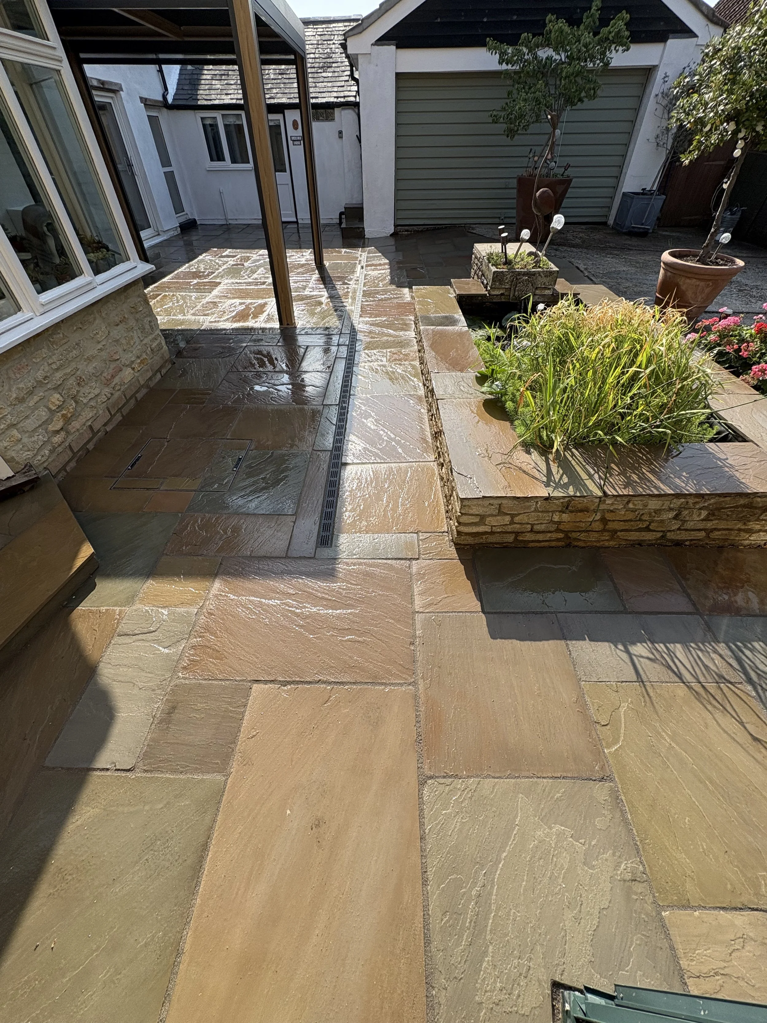 A patio with wet stone tiles, a raised flower bed with plants, potted trees, and a garage door in the background.