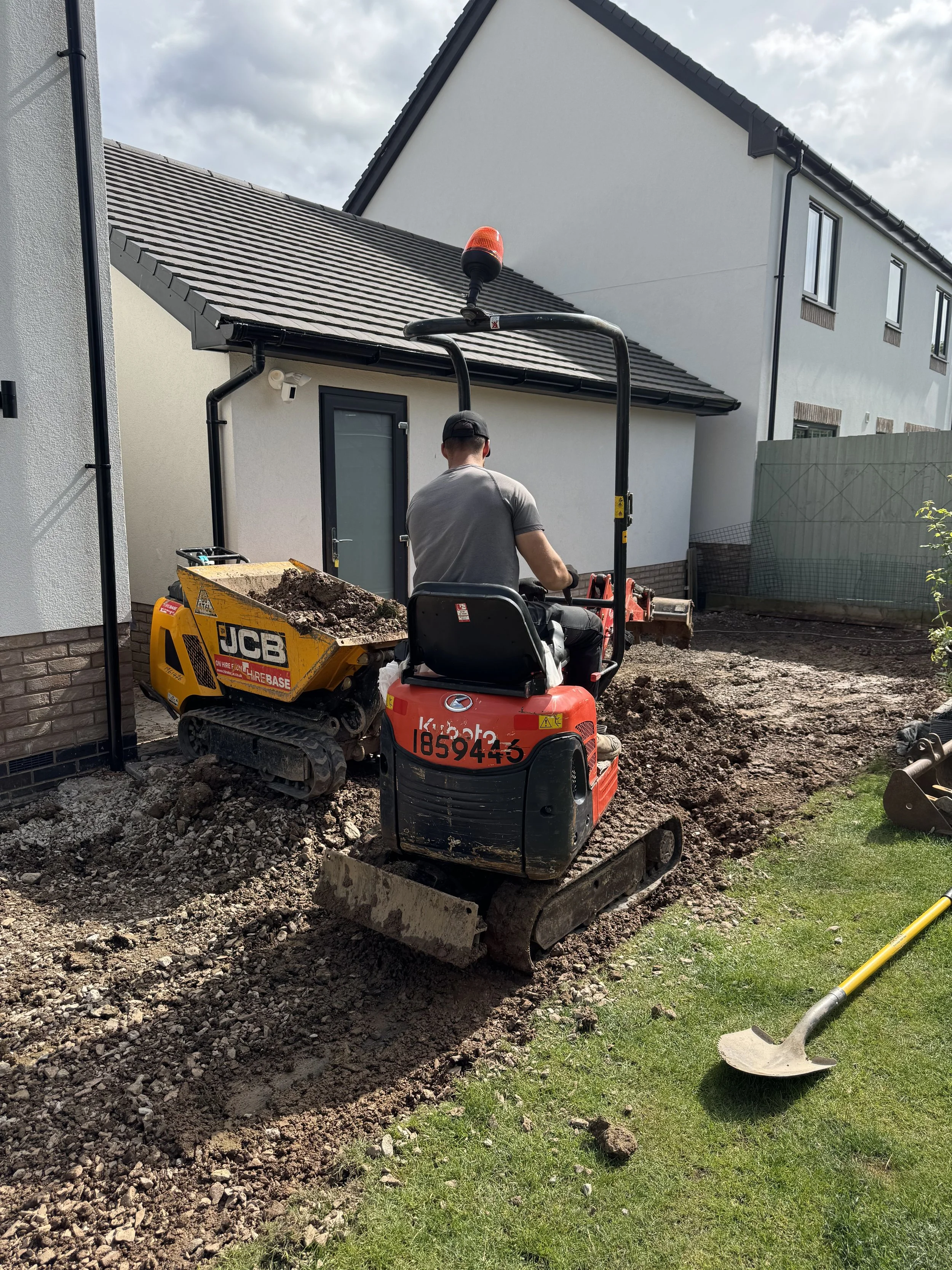Person operating a small excavator on a backyard construction site, digging soil near a white house with a gray roof.