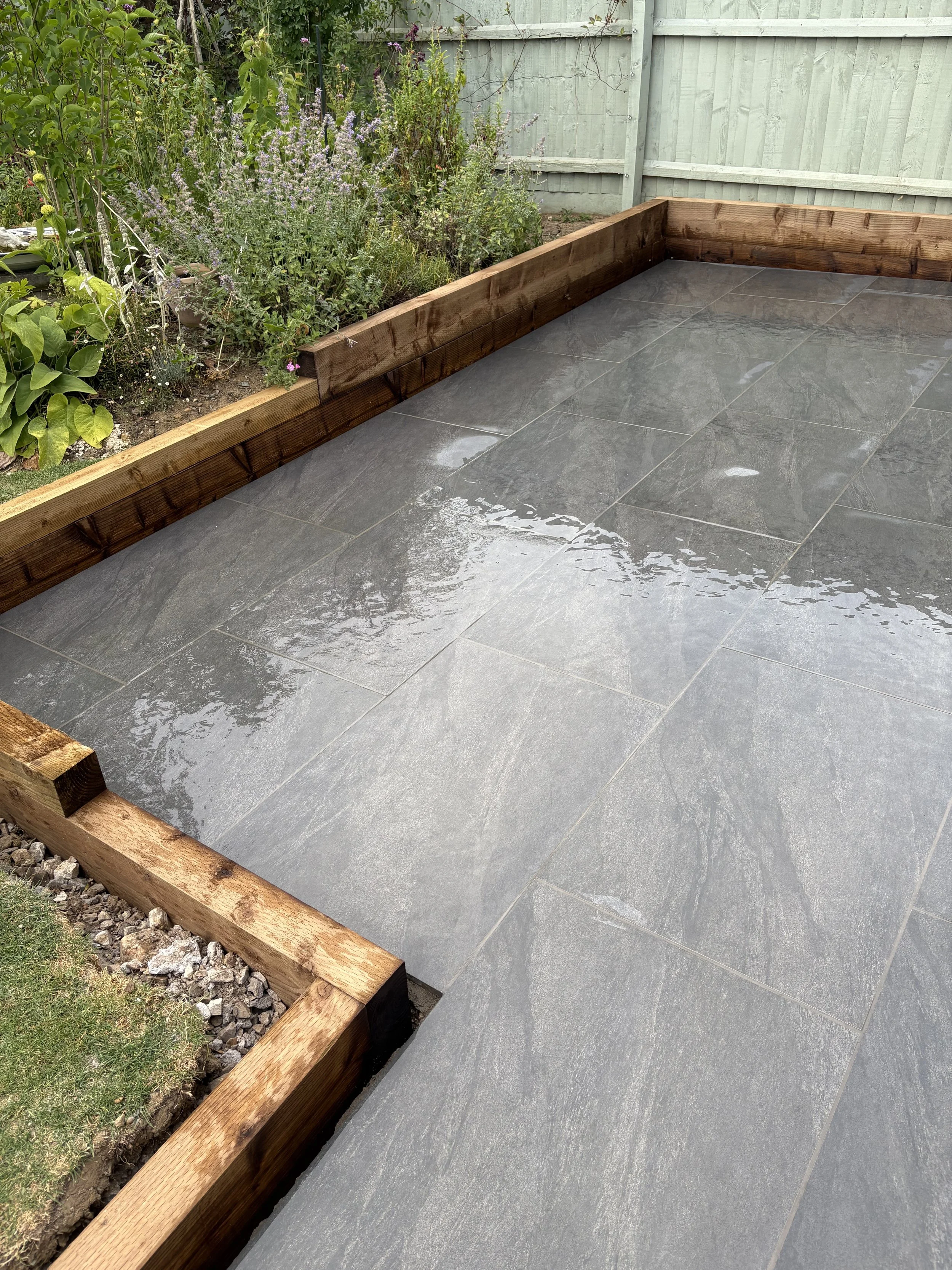 A wet gray tiled patio area with wooden edging, next to a garden bed with various plants and flowers, and a light green wooden fence in the background.