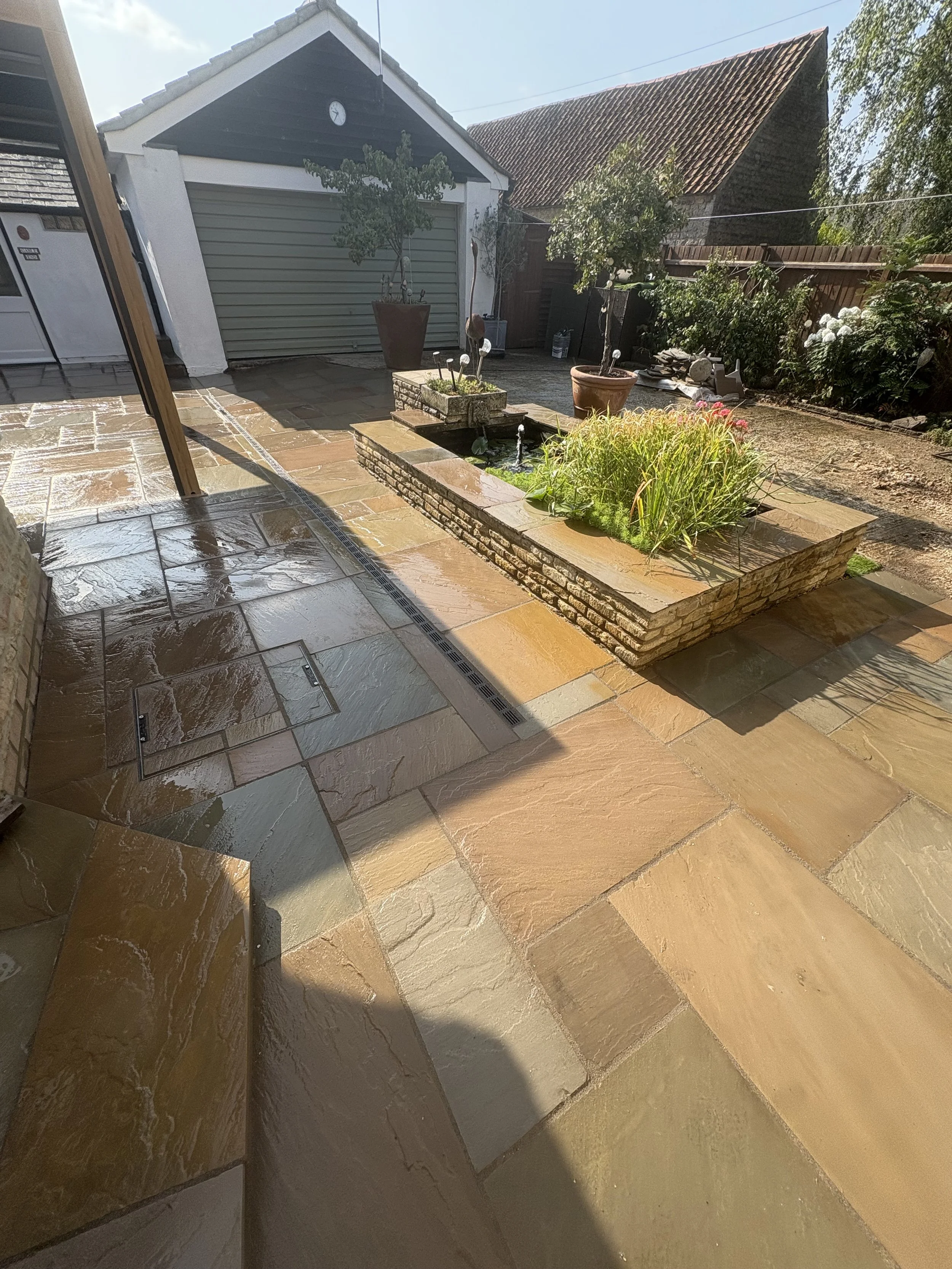 Sunlit backyard patio with stone flooring, a raised brick garden bed with plants and a small fountain, surrounded by potted plants, trees, and a shed with a closed garage door.