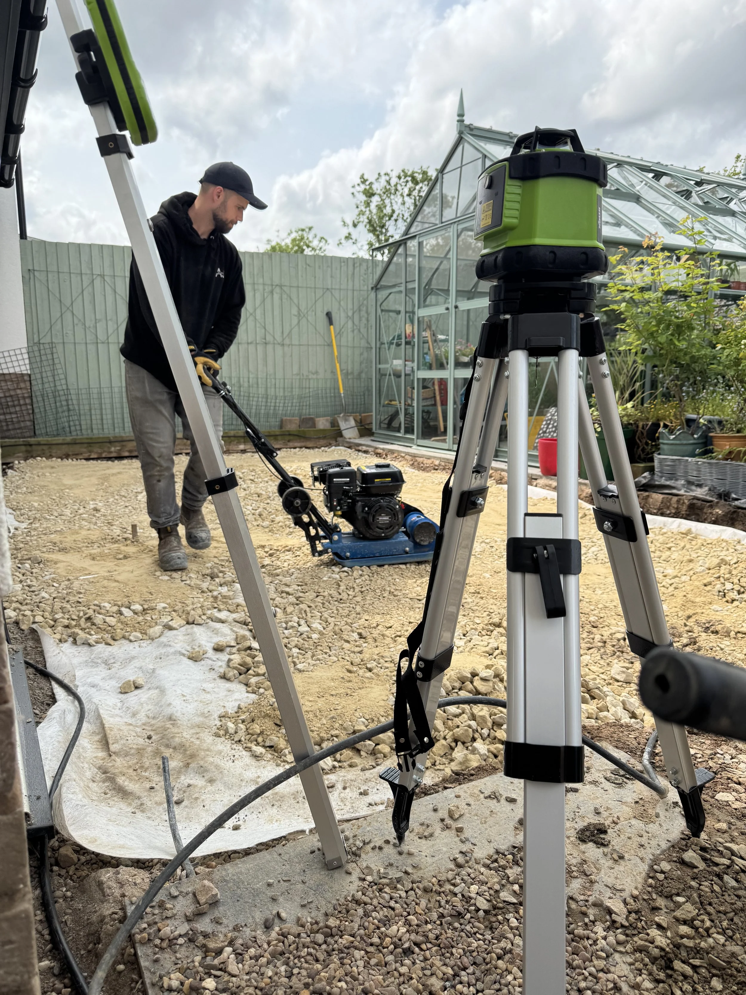 A person working on a construction site with surveying equipment, including a tripod with a green device, and a compact excavator, near a greenhouse with plants outdoors.