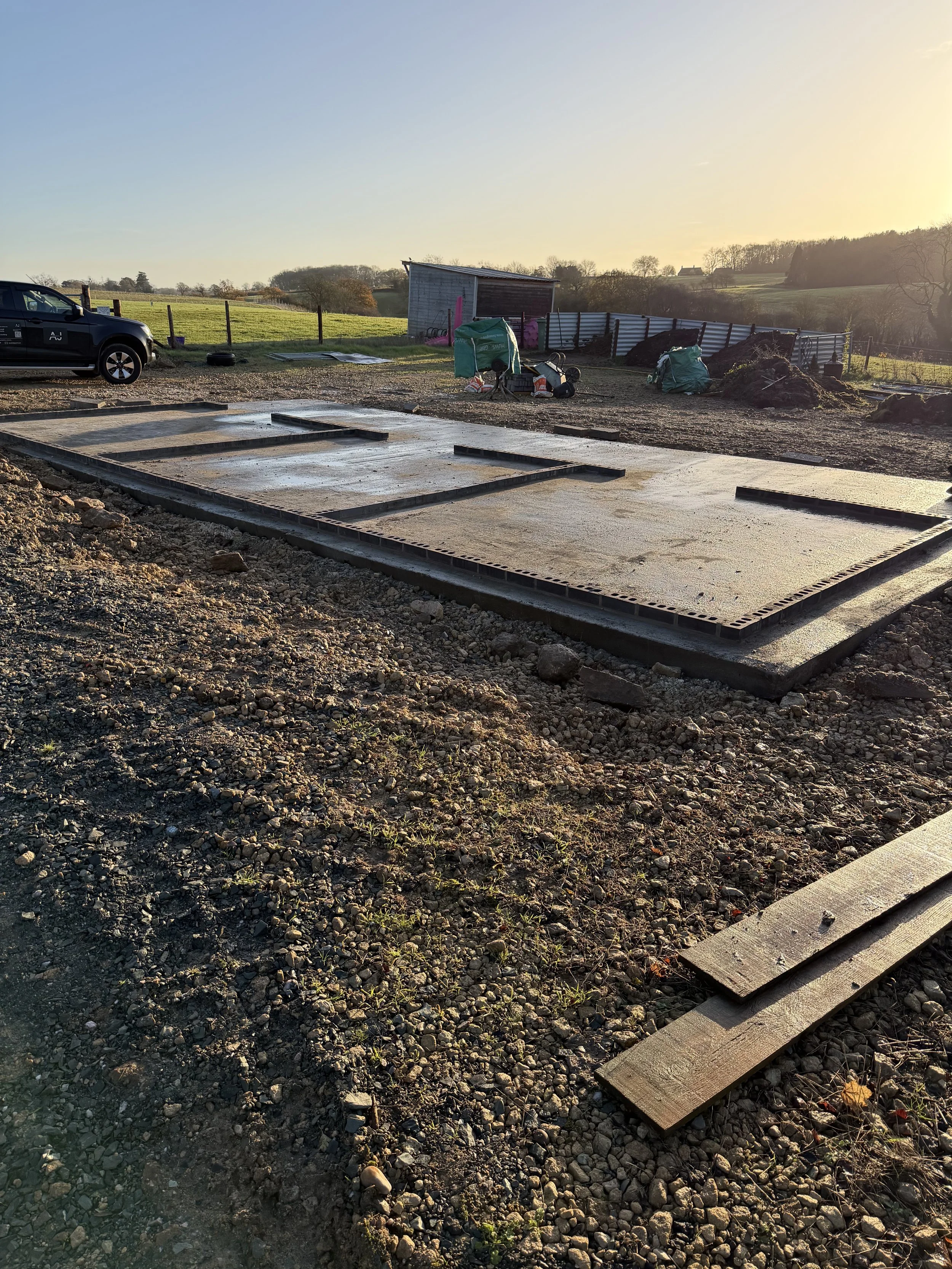 Construction site with concrete foundation slab, wooden planks, a parked black vehicle, and construction tools and supplies, with a rural landscape in the background during sunset.