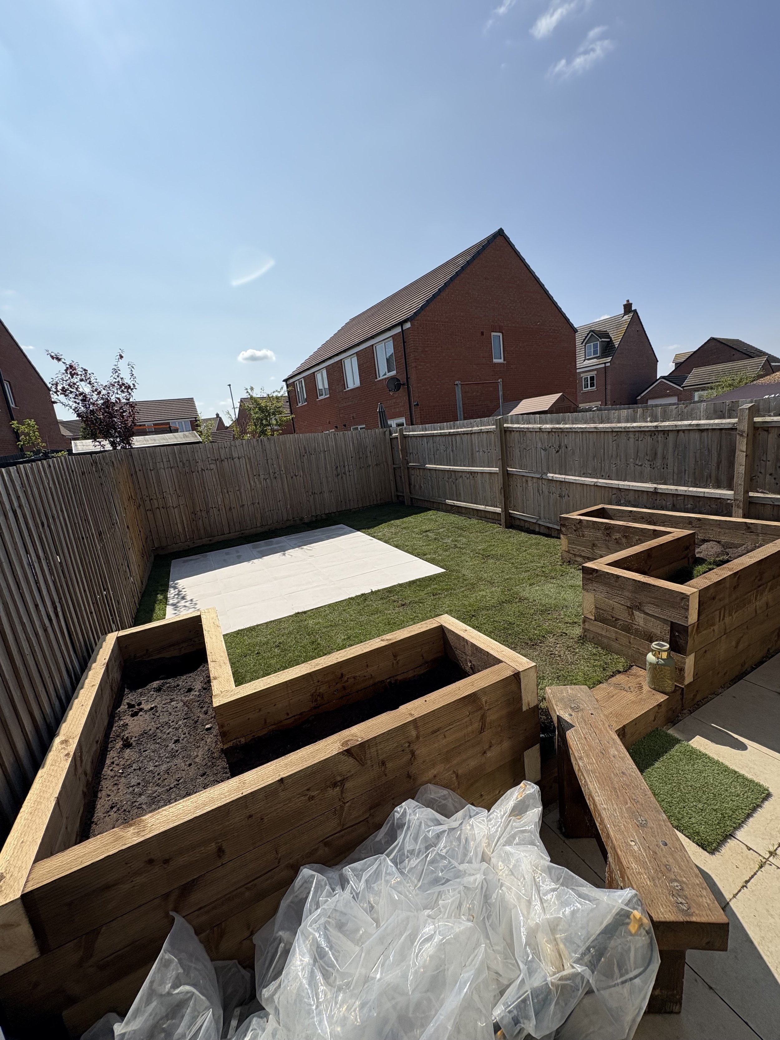 Backyard garden with wooden raised beds, some filled with soil, and a grassy area with a white tarp, surrounded by a wooden fence, on a sunny day.