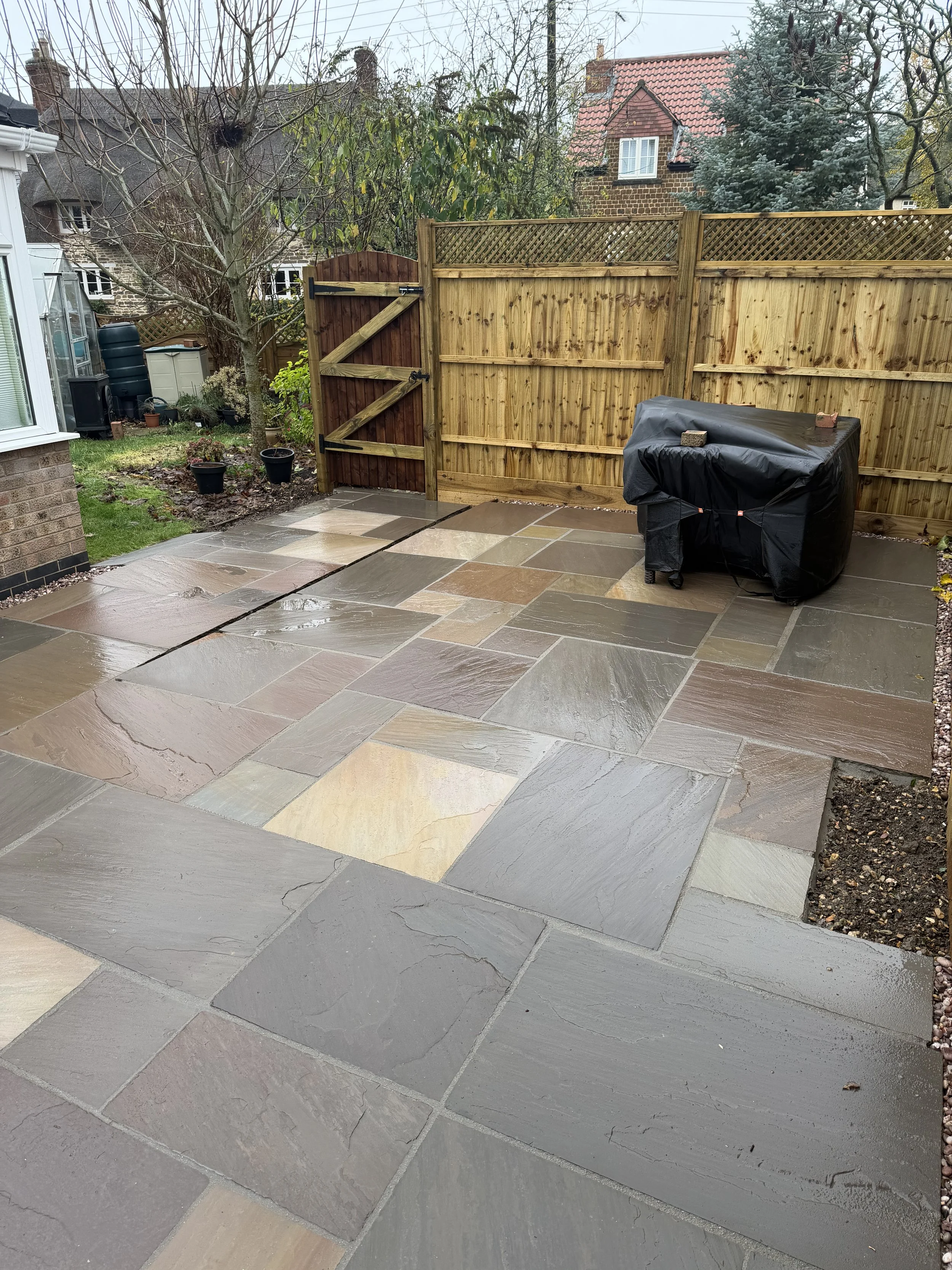 Wet stone patio in a backyard enclosed by a wooden fence, with a barbecue covered in a black protective cover on the right, and some small potted plants near a tree on the left, with neighboring houses in the background.