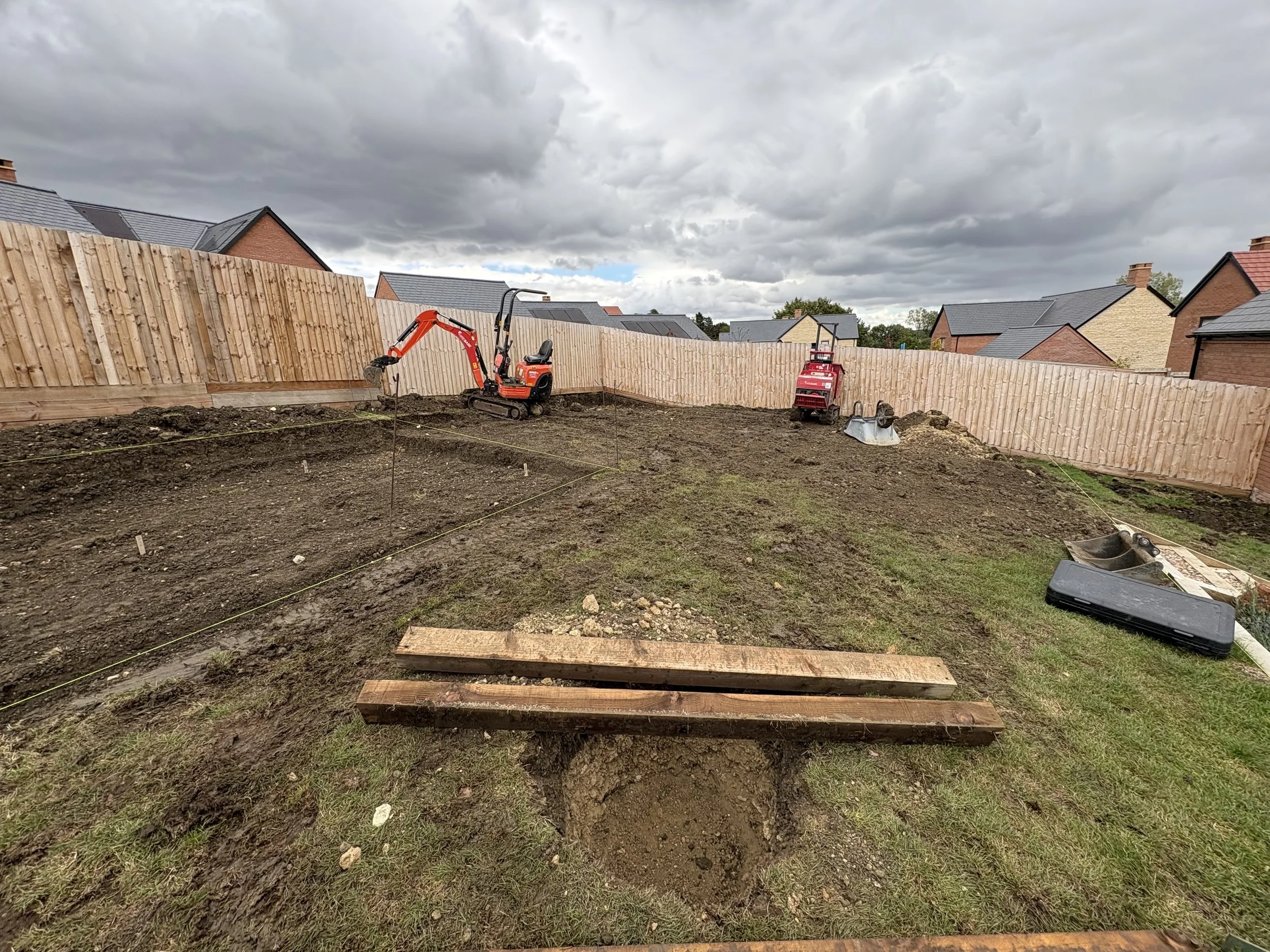 Construction site in backyard with dirt, wooden planks, mini excavator, small roller compactor, and other construction tools, enclosed by a wooden fence, under a cloudy sky.