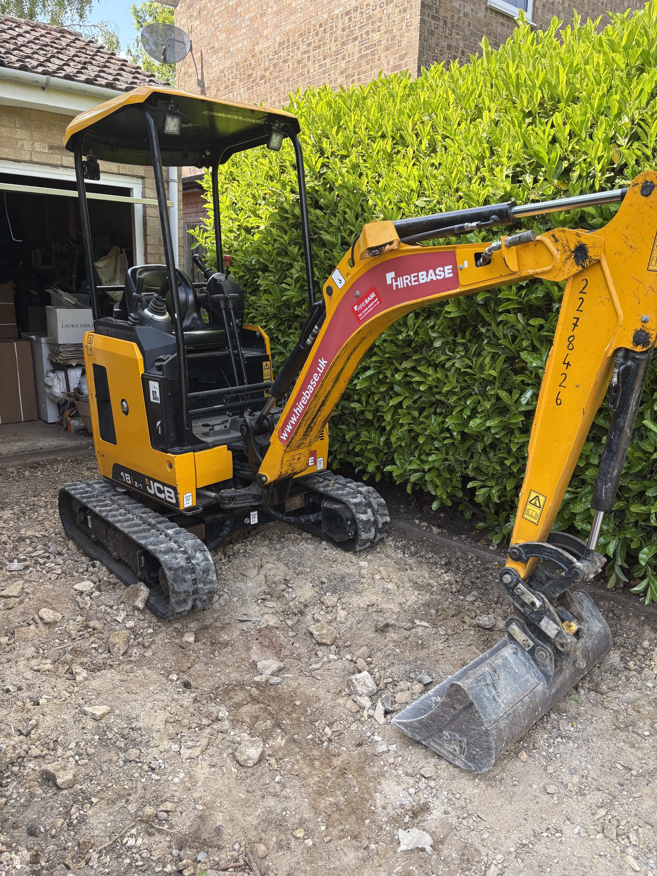 A small yellow JCB mini excavator with black tracks parked on dirt in a residential yard, next to a green hedge and a brick house.