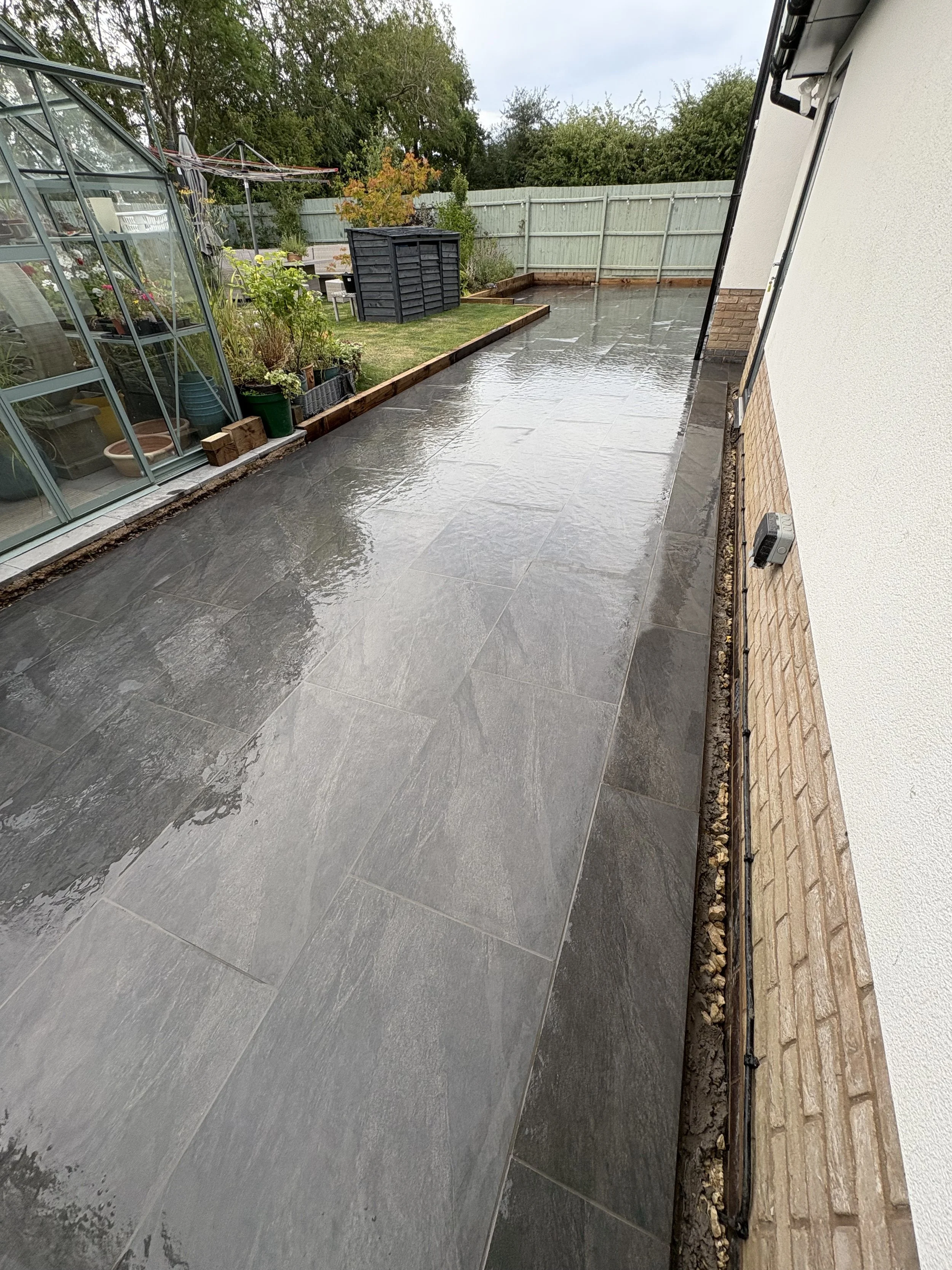 Wet outdoor patio with large gray tiles, garden area with plants and shrubbery, and a fenced backyard.