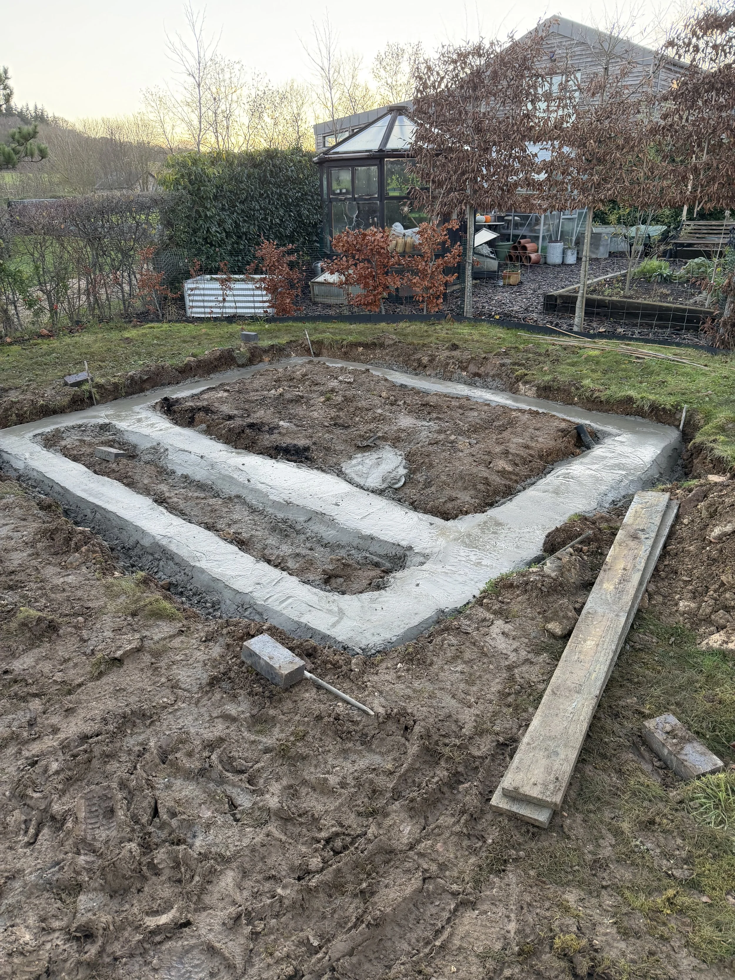 Construction site in a backyard with a rectangular foundation outline and concrete footing, surrounded by dirt and construction tools, with garden sheds and trees in the background.