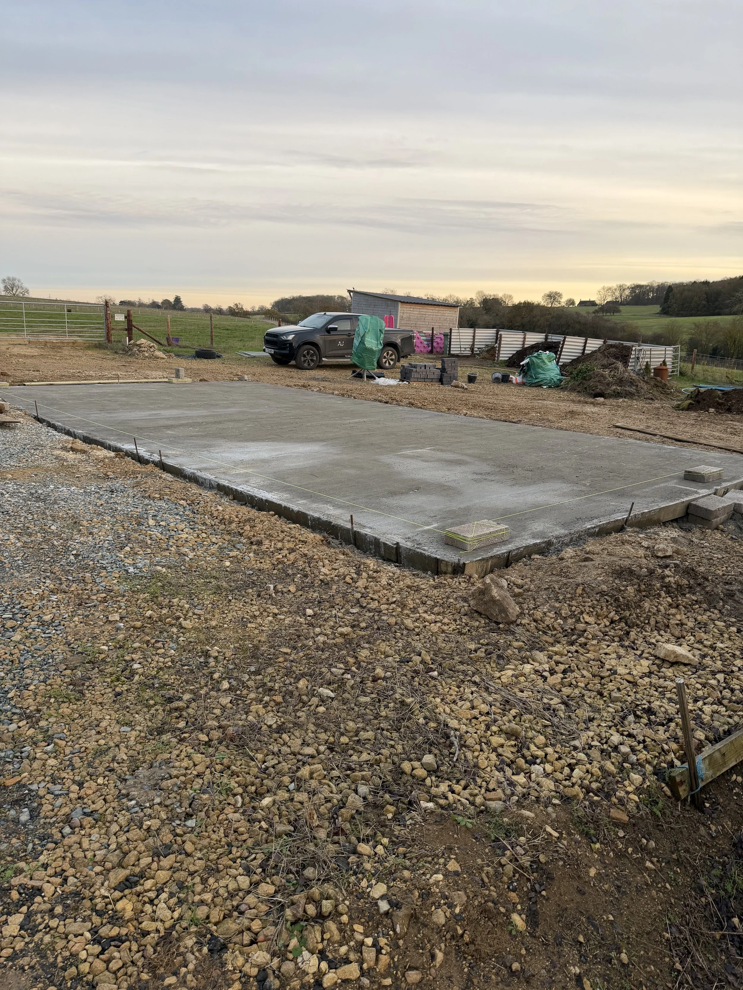 Freshly poured concrete slab foundation for construction with wooden formwork, construction tools, and materials in a rural outdoor setting.