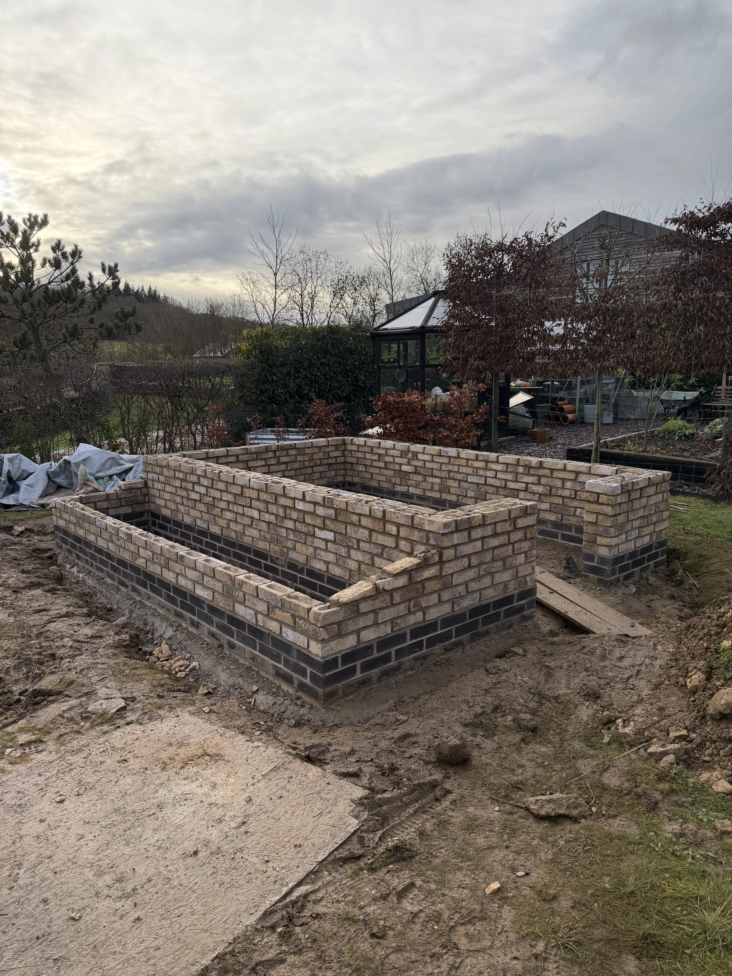 A brick framework for a garden bed under construction in a backyard, with a house, trees, and a greenhouse in the background.