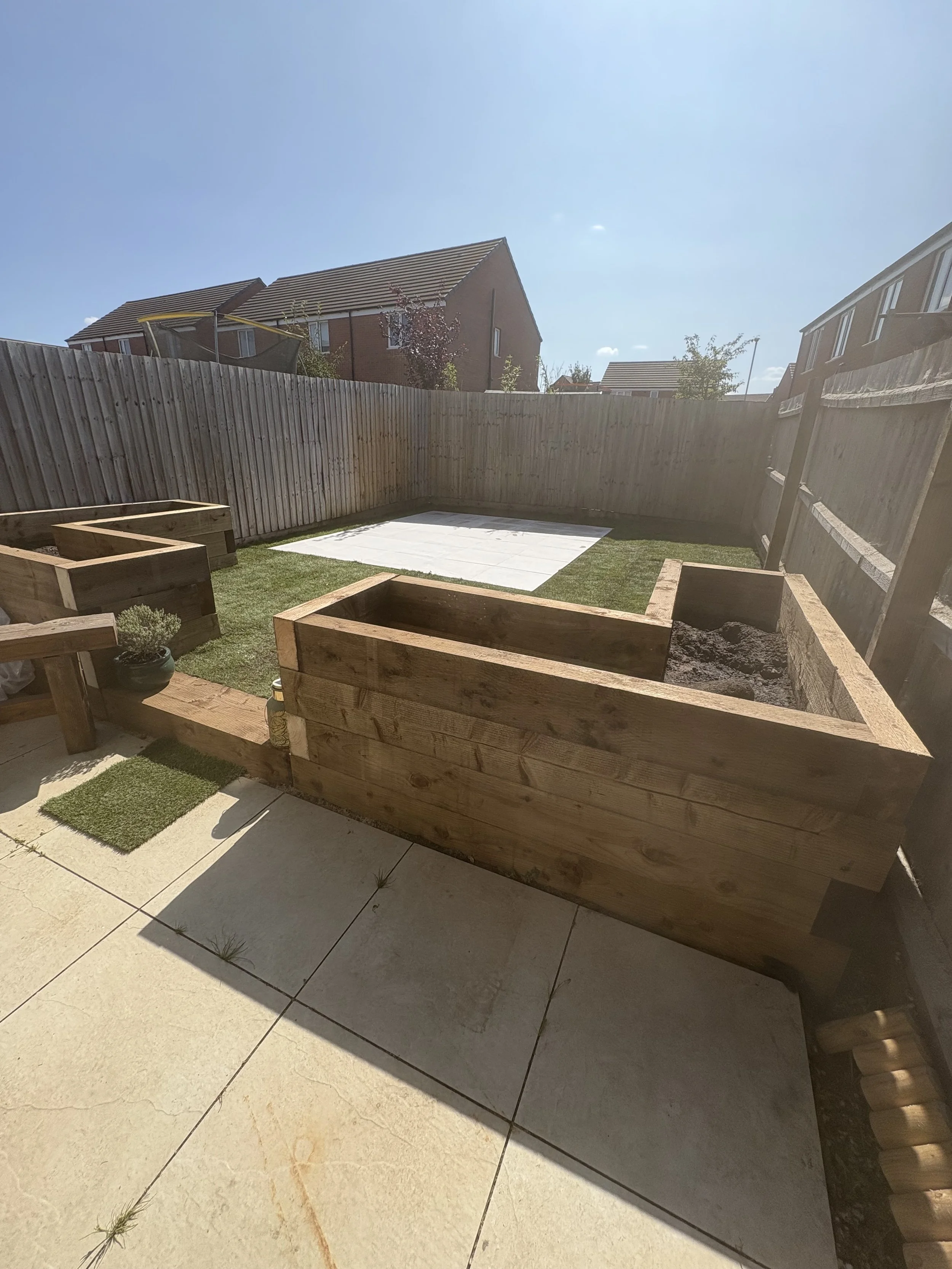 Backyard with wooden raised garden beds, a small patch of grass, a tiled area, and a wooden fence under a clear blue sky.