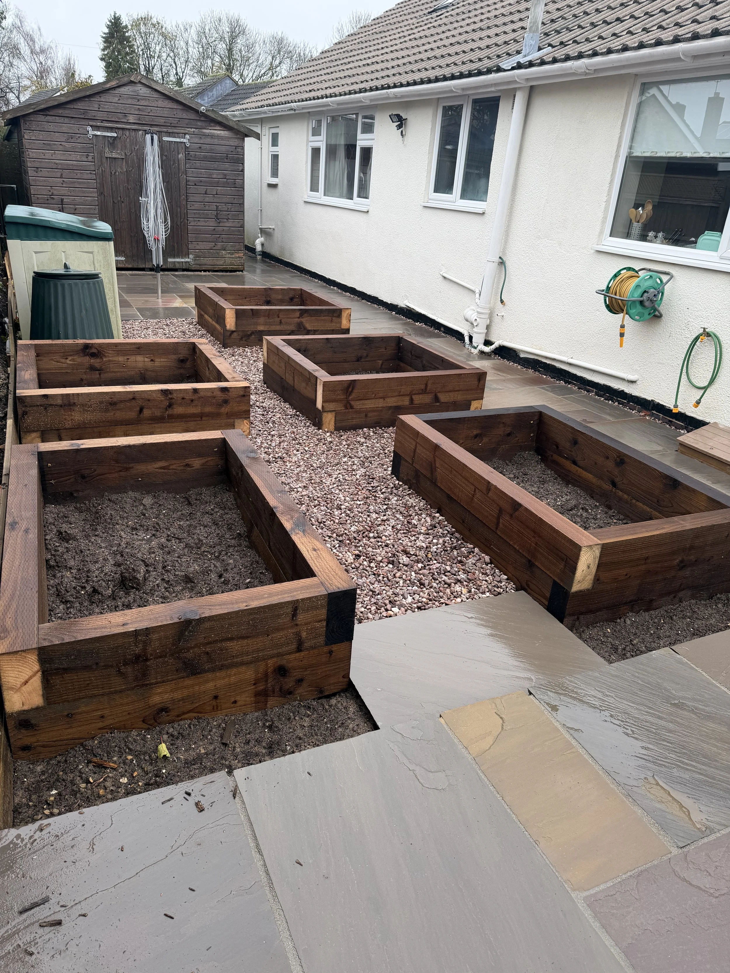 Backyard with six wooden garden beds filled with soil, set on a stone patio, with a shed and house in the background.