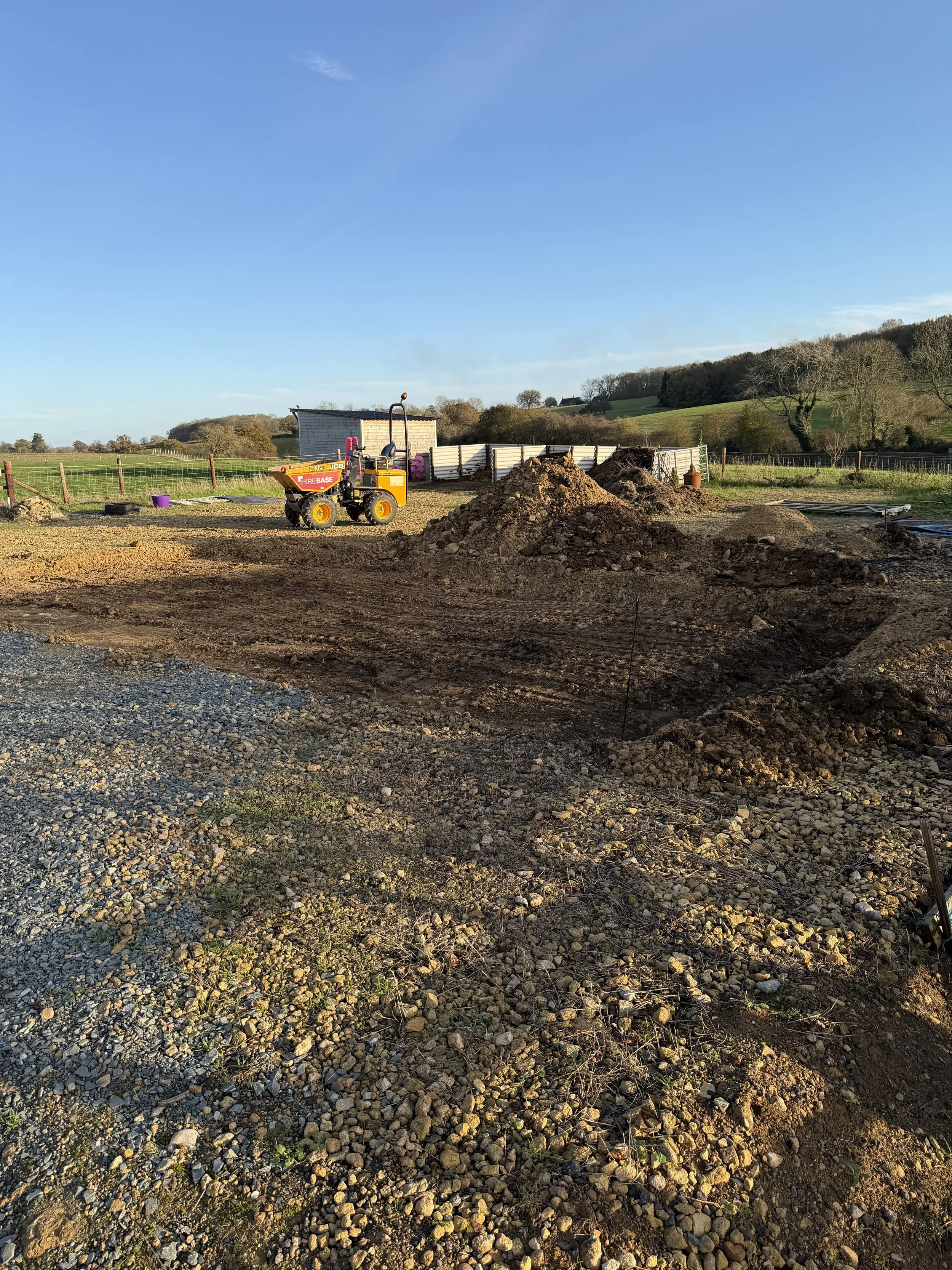 Construction site with a small yellow and purple roller compactor and piles of dirt, fencing, and open fields in the background.
