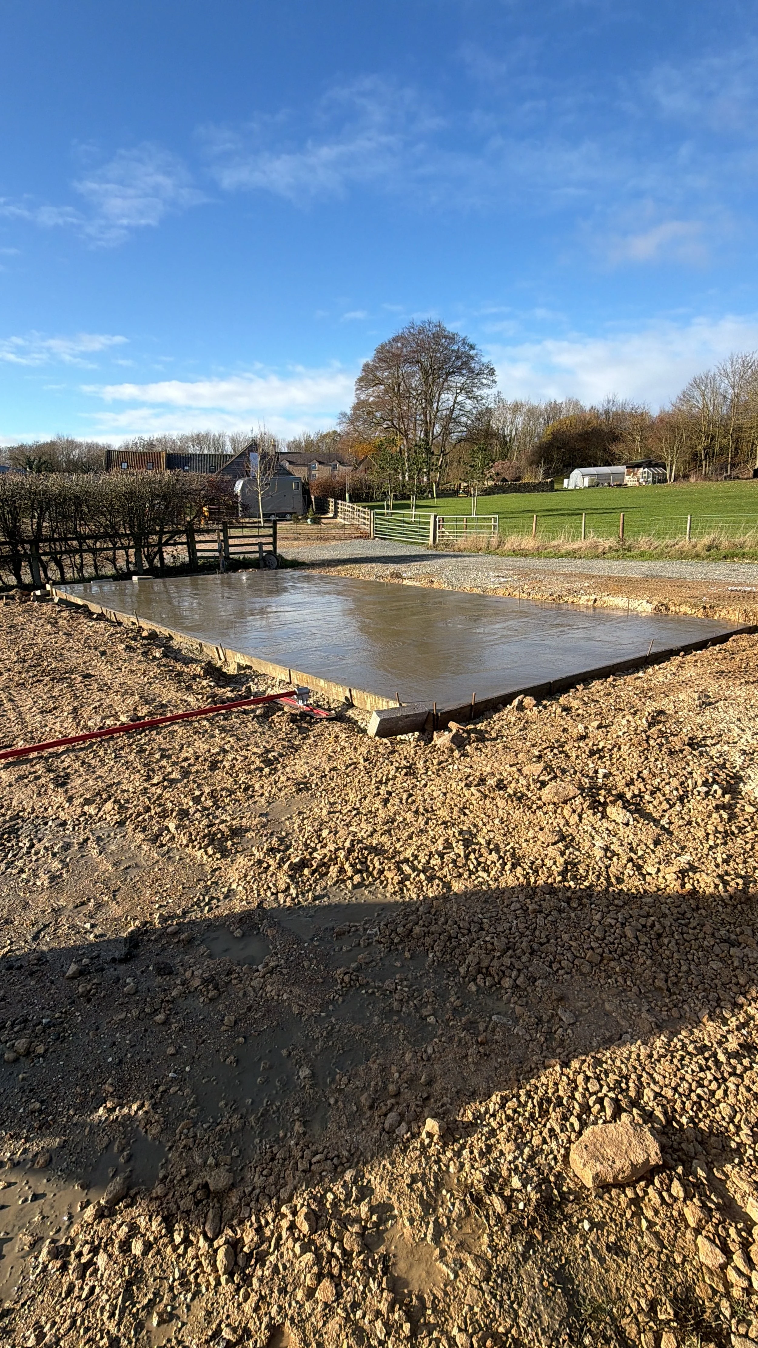 Freshly poured concrete slab on a construction site in a rural area with trees, green fields, and blue sky in the background.