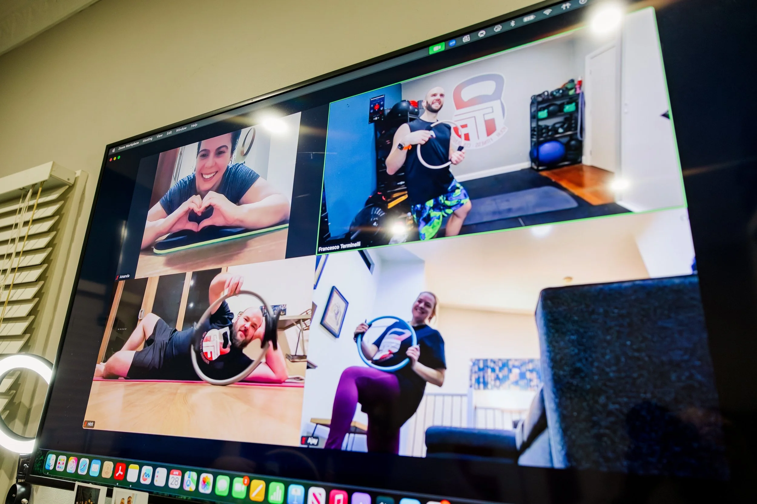 Man lying on exercise mat with one leg raised, doing a workout at home gym with shelves of weights and fitness equipment in background, participating in virtual workout on laptop.