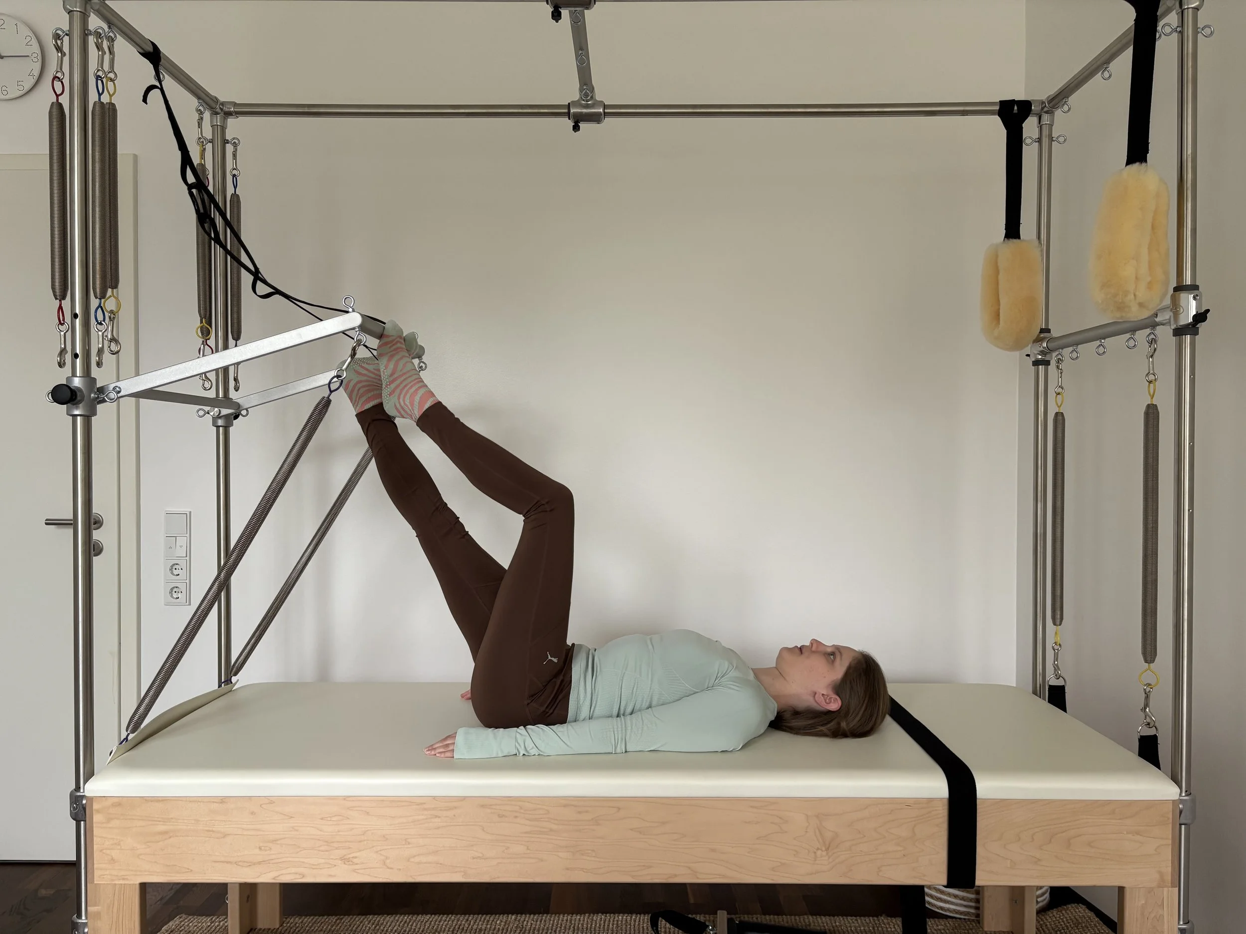 Melanie Ott liegt auf dem klassischen Pilatesgerät Cadillac. Die Füße sind auf der TowerBar aufgestellt. Sie demonstriert die Übung Footwork.