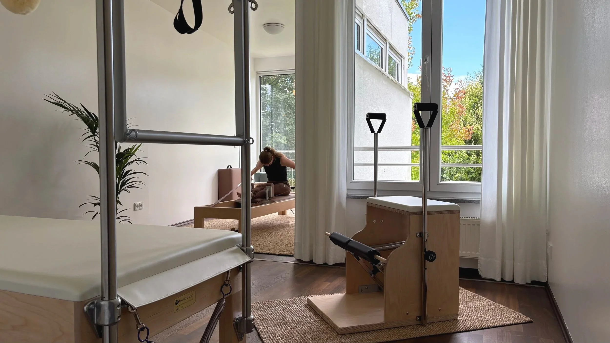 Studioraum mit traditionellen Pilatesgeräten und einer Person, die auf einem Reformer sitzt, in einem hellen Raum mit großen Fenstern und Blick in die grüne Natur.