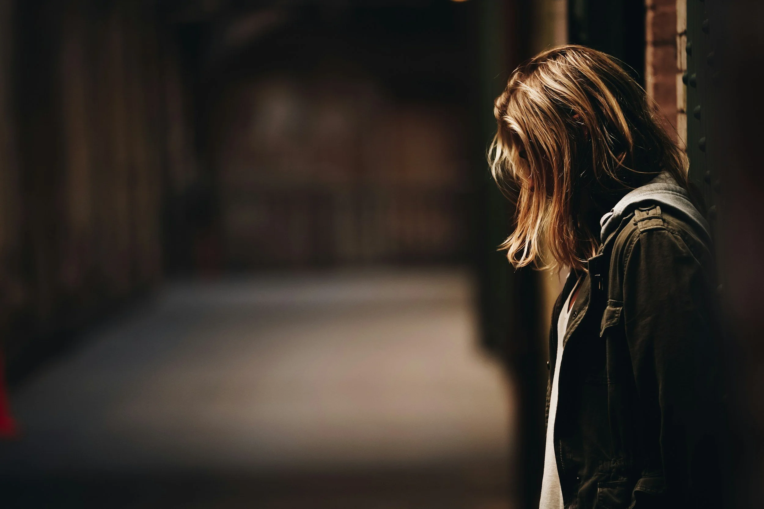 A woman with long hair, dressed in a dark jacket, leaning against a brick wall in a dimly lit alleyway.