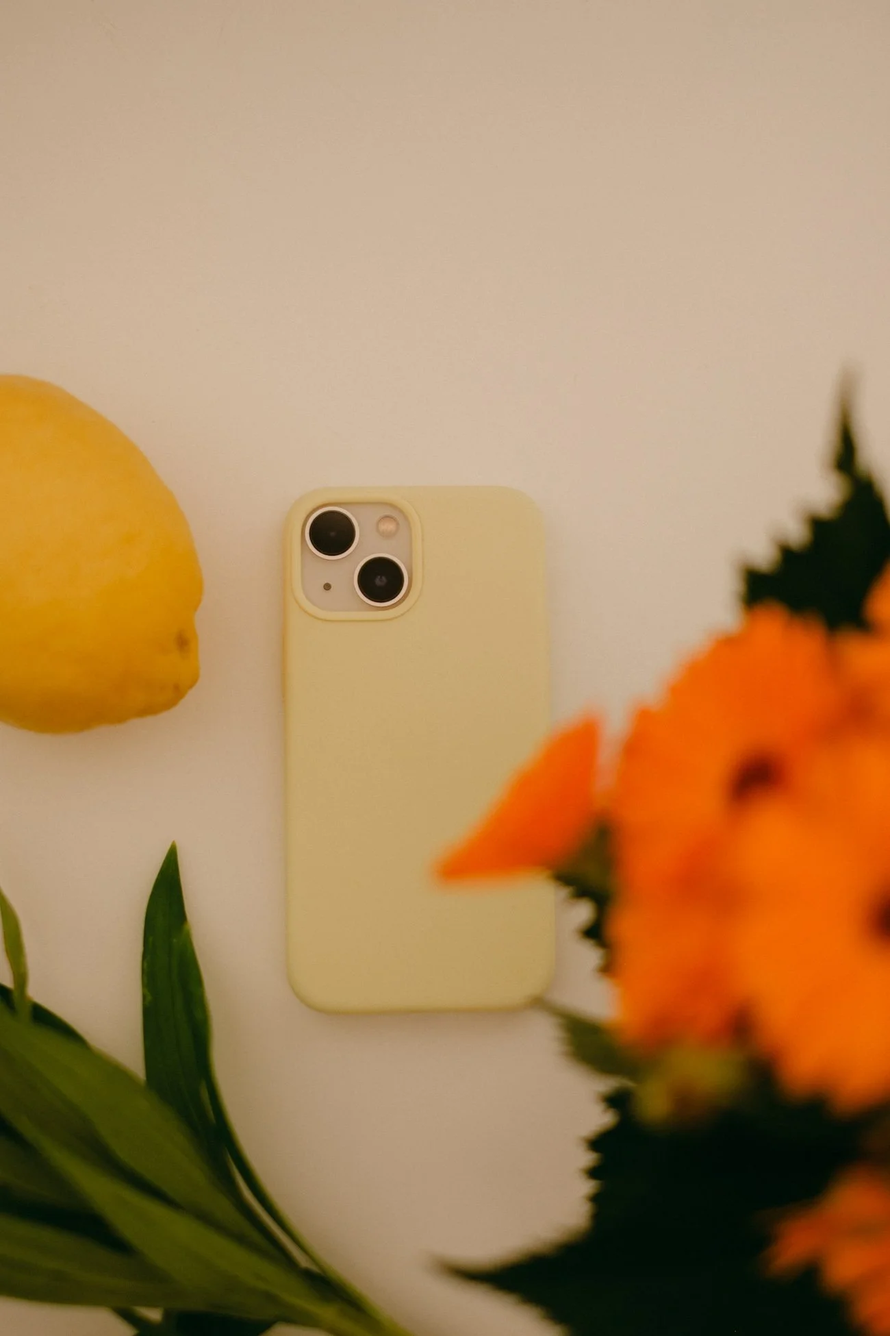 Uno smartphone giallo chiaro è posato su una superficie bianca, circondato da un limone a sinistra e alcuni fiori arancioni e verdi in primo piano a destra.