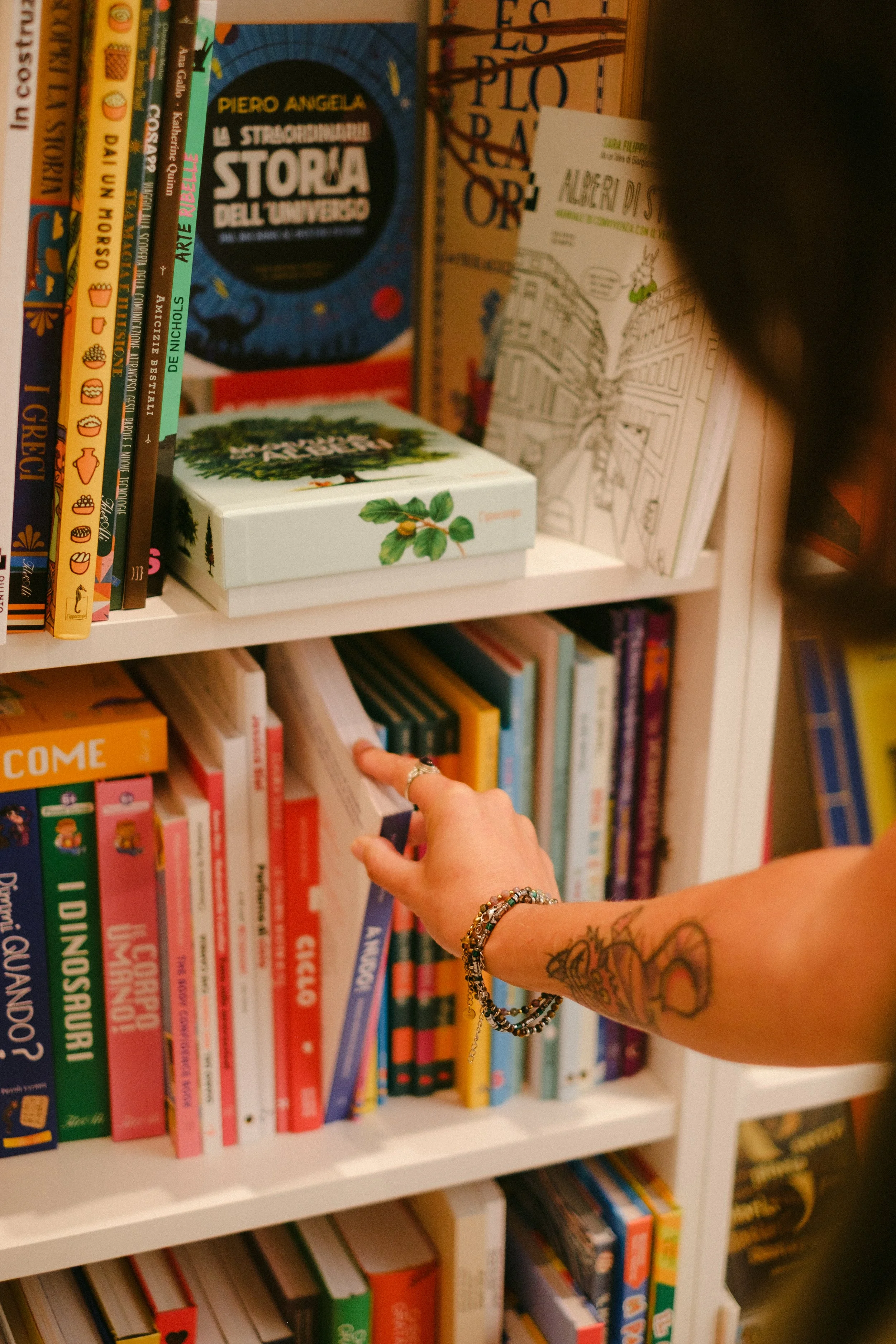 Una persona che sfoglia un libro su una libreria piena di libri colorati e varia, con braccialetti e tatuaggi visibili sul braccio.