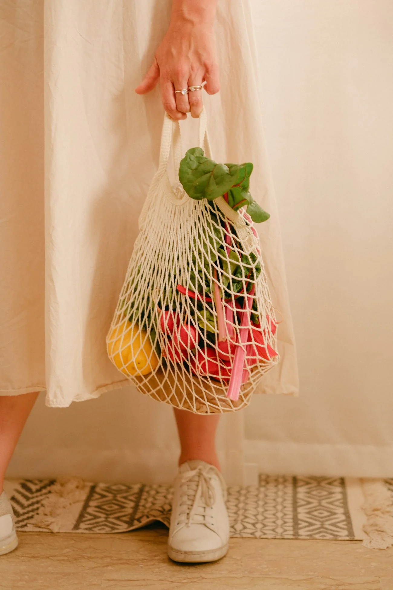 Una persona tiene un sacco di tela con frutta e verdura, come limoni, pomodori, e foglie verdi, indossando scarpe sportive bianche e una gonna chiara.