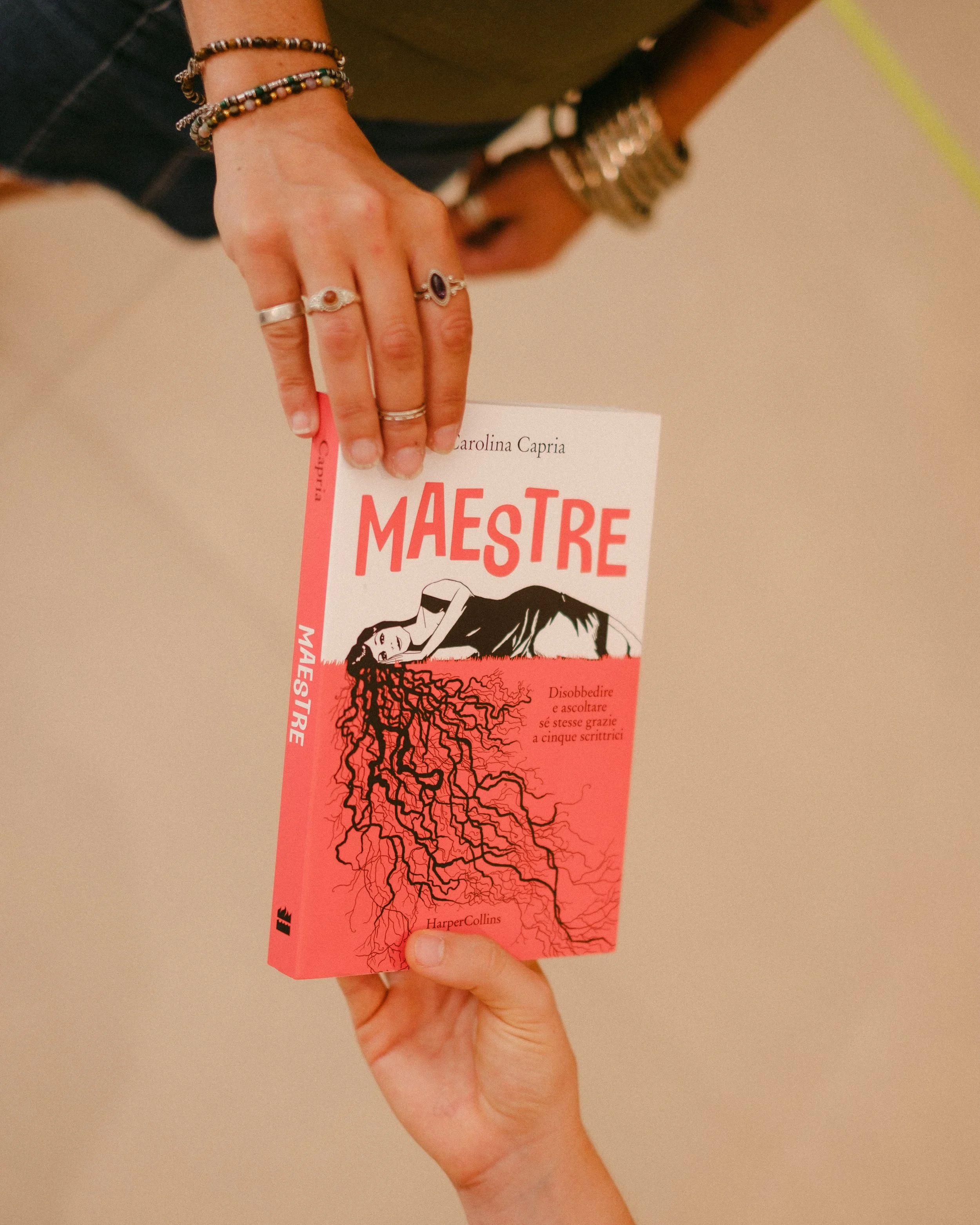 Due mani che si scambiano un libro intitolato "Maestre" con una copertina rossa e un'illustrazione di una donna sdraiata con capelli che si diramano come radici.
