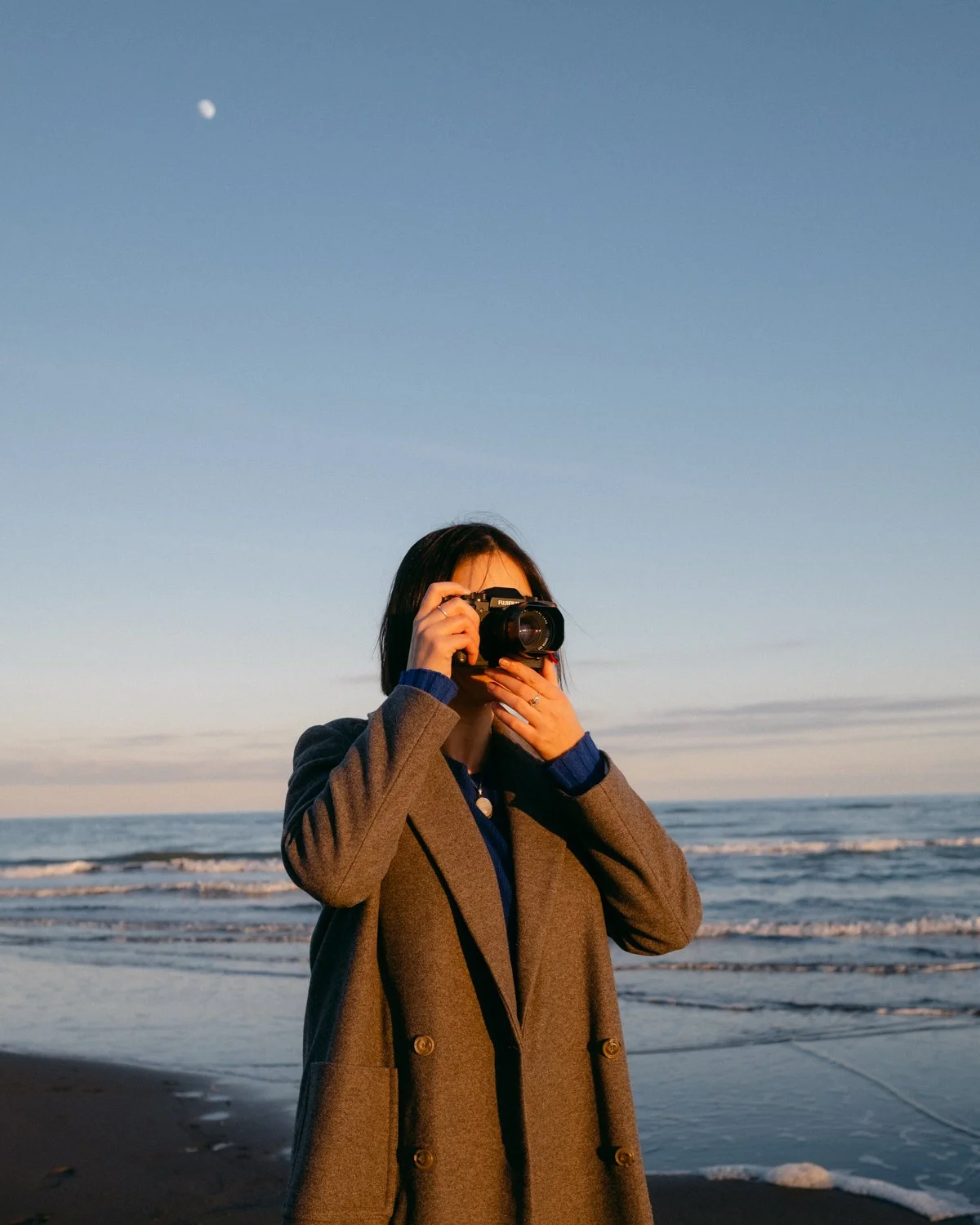 Donna con cappotto grigio che scatta foto sulla spiaggia al tramonto con il cielo al tramonto e la luna visibile