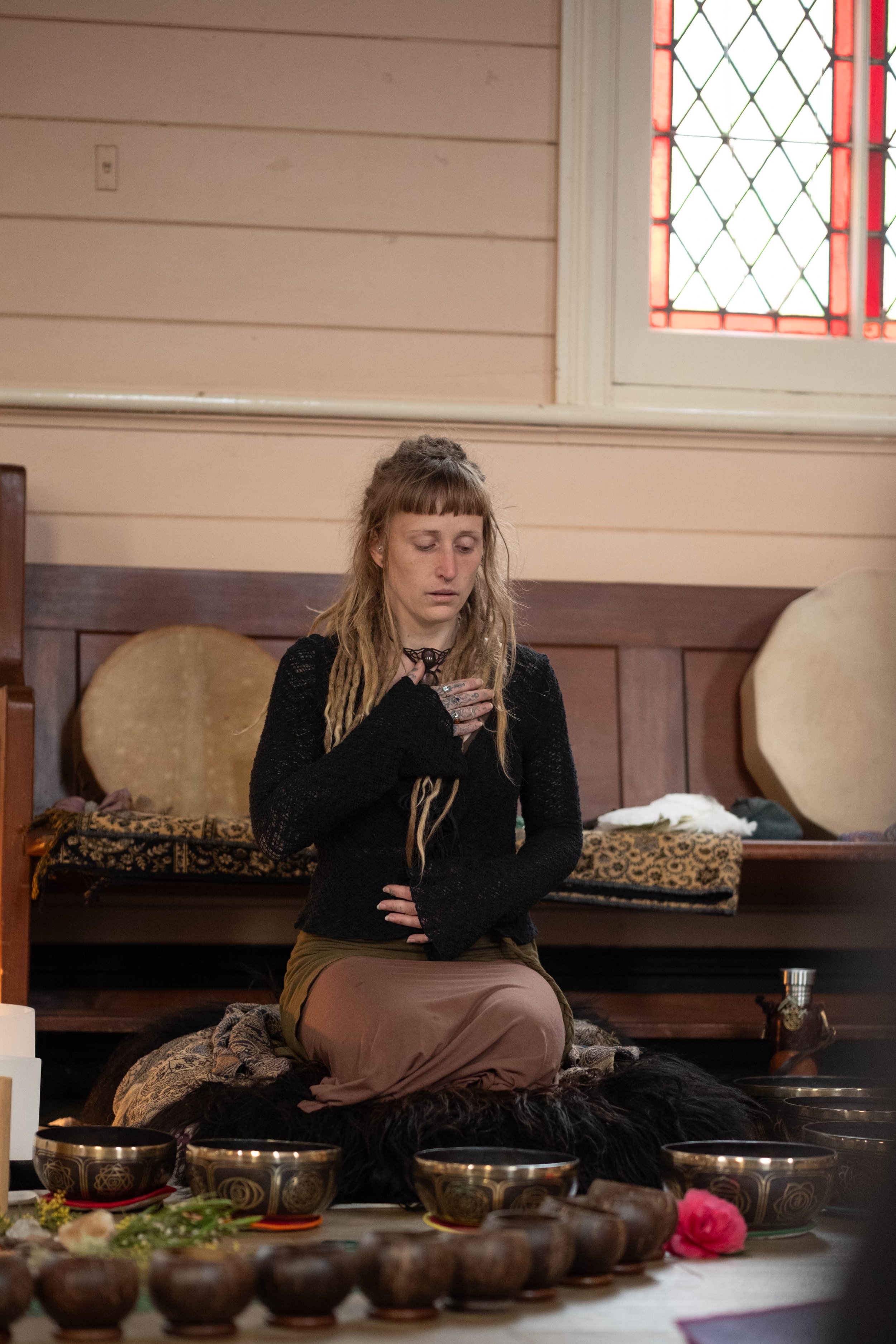 A woman with long dreadlocks kneeling on a black fur rug, with her hand over her chest and eyes closed, surrounded by bowls and flowers, inside a room with wooden walls and a stained glass window.