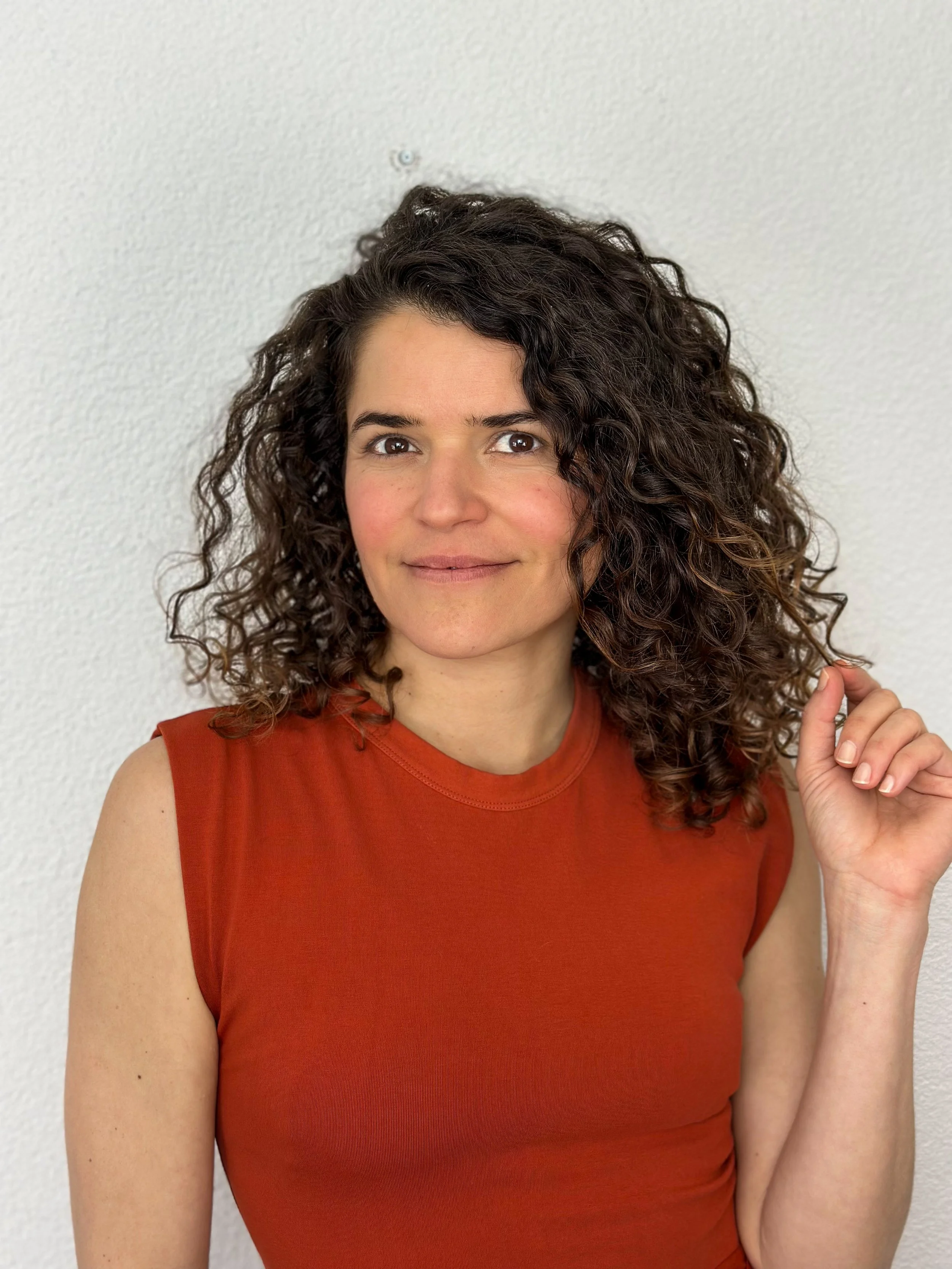 Eine Frau mit lockigem Haar und einem orangefarbenen ärmellosen Oberteil steht vor einem neutralen Hintergrund. Unternehmerin Ludmila De Albuquerque Lins.