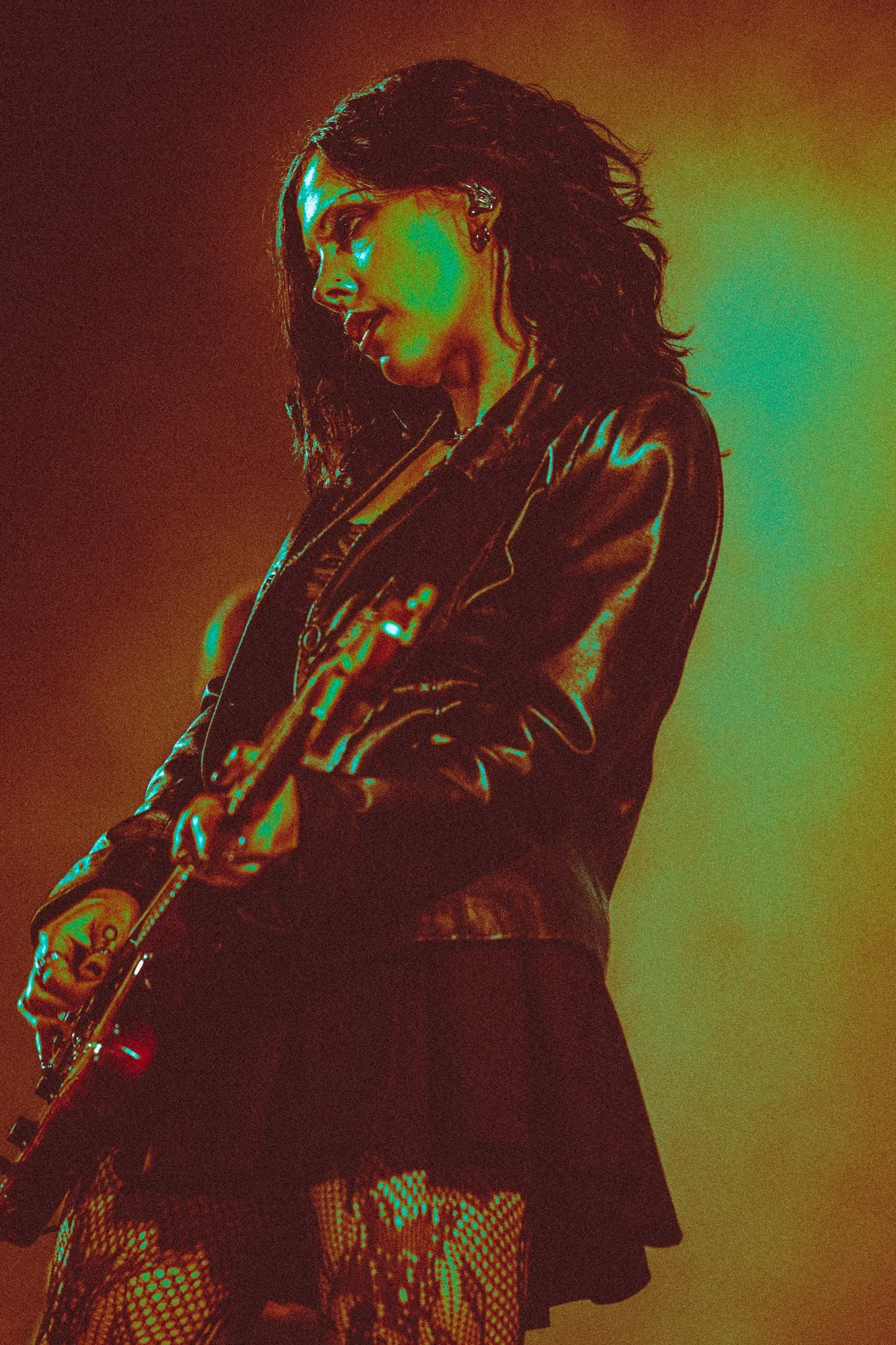 A person with long, curly hair dressed in a shiny black leather jacket playing a guitar on stage with colourful lights.