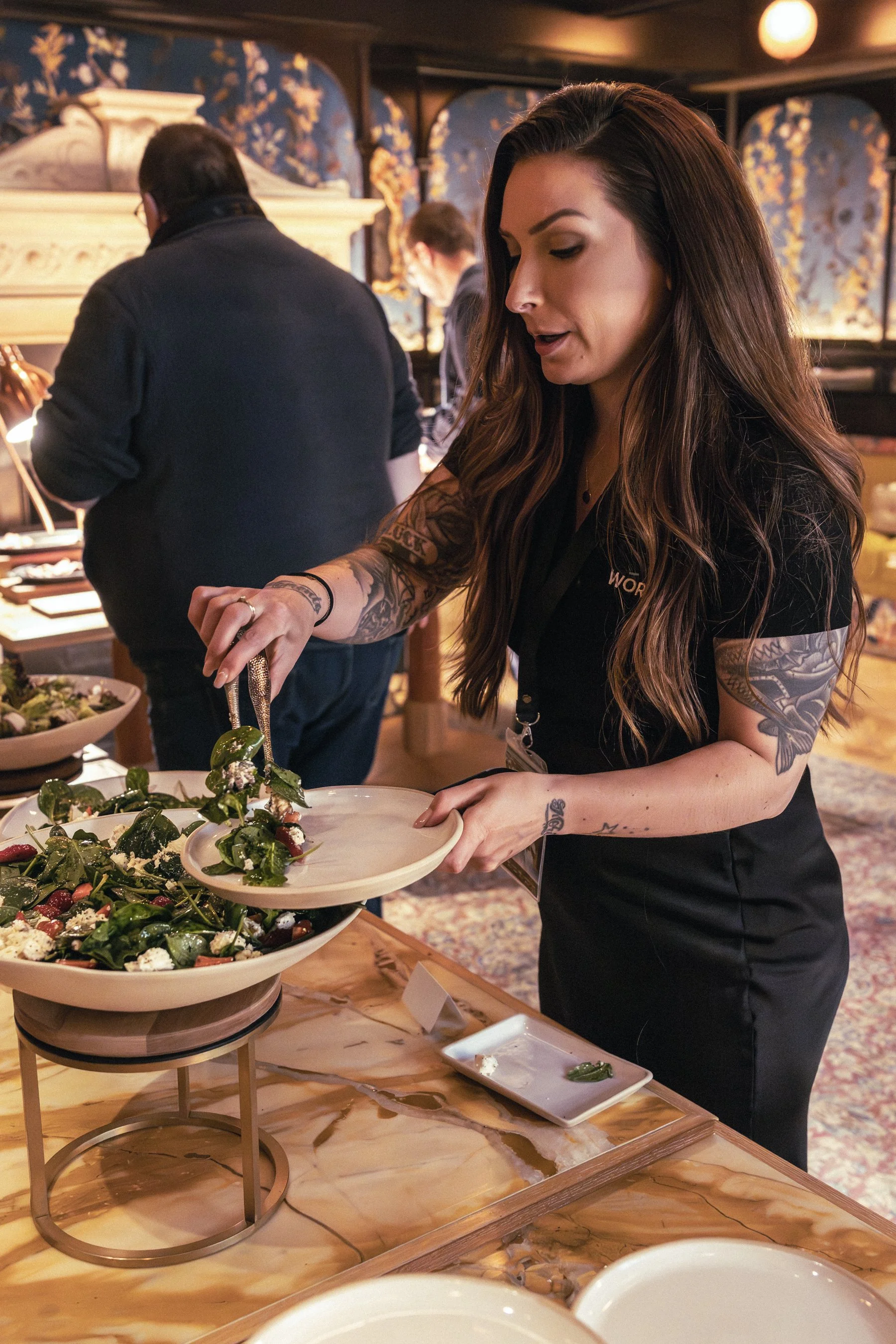 A woman with long brown hair and tattoos on her arms is serving herself a salad from a buffet table at a restaurant, with other people in the background.