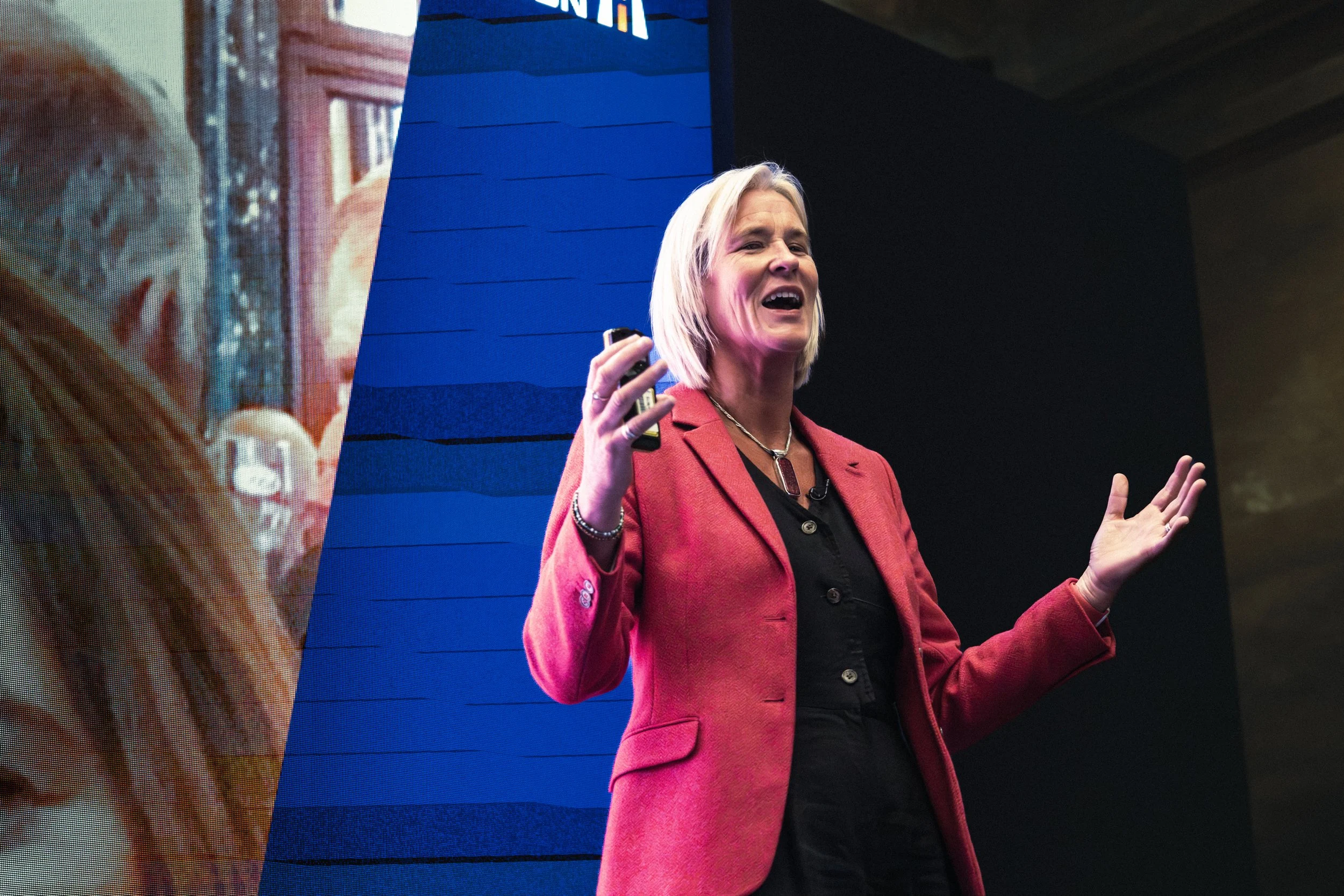 A woman with blonde hair wearing a red blazer giving a presentation on stage. She is speaking and gesturing with her hands, with a large screen or display behind her.