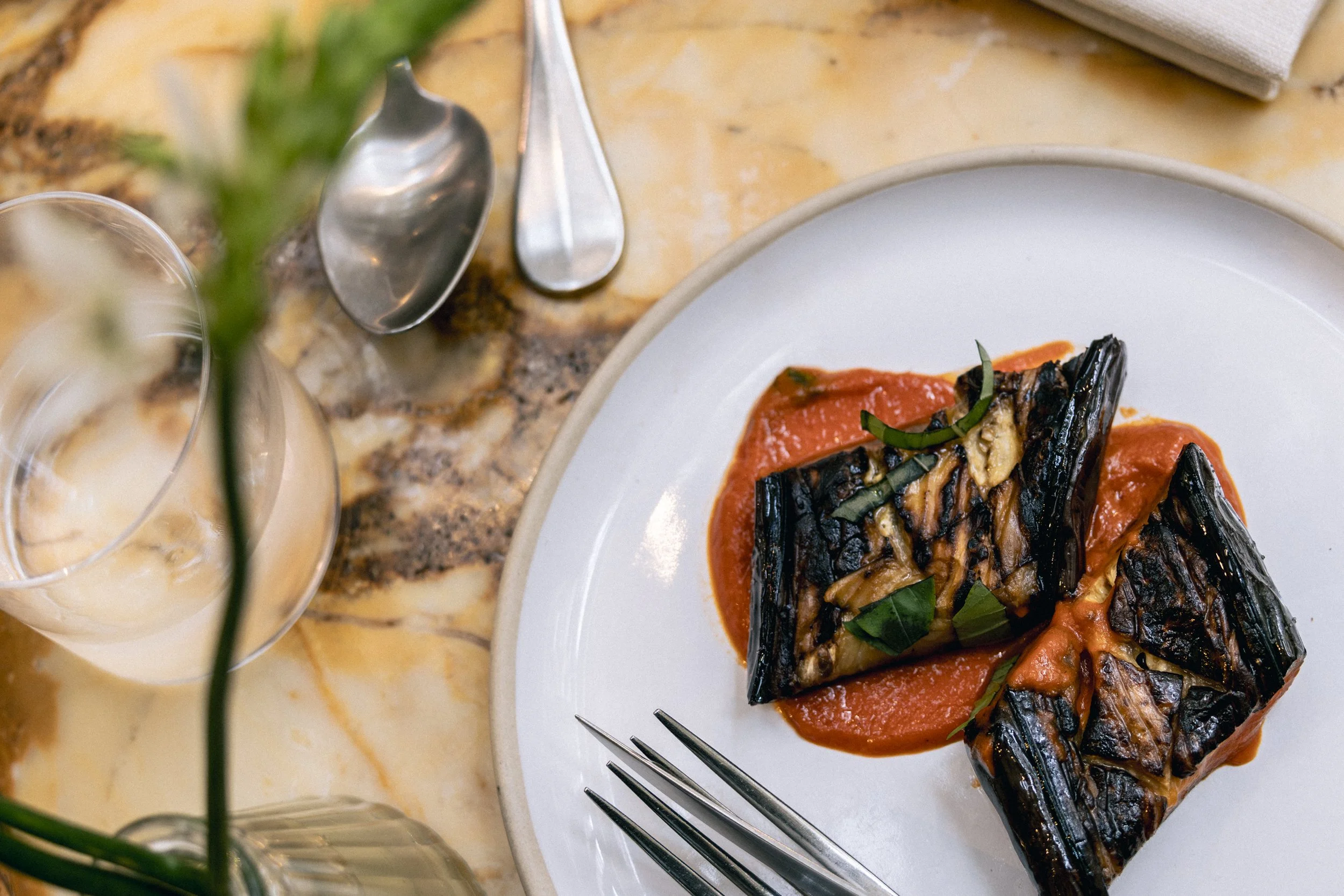 A plate of grilled fish topped with tomato sauce and garnished with chopped green herbs, with a fork resting on the plate. In the background, there are two spoons and a glass of beverage with ice, positioned on a countertop, along with a folded napki