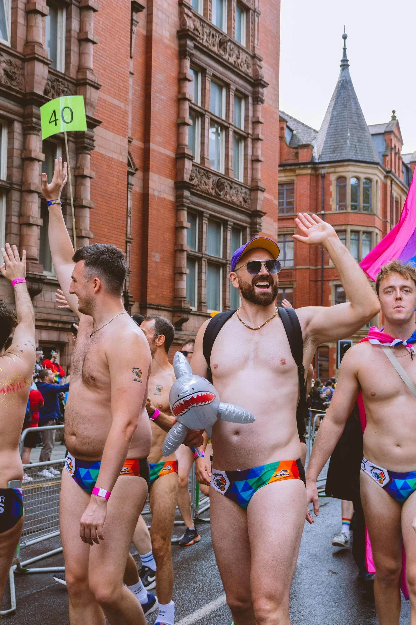 Group of shirtless men wearing rainbow-themed underwear participating in a parade, with some holding pink and purple flags, in front of a red brick building.