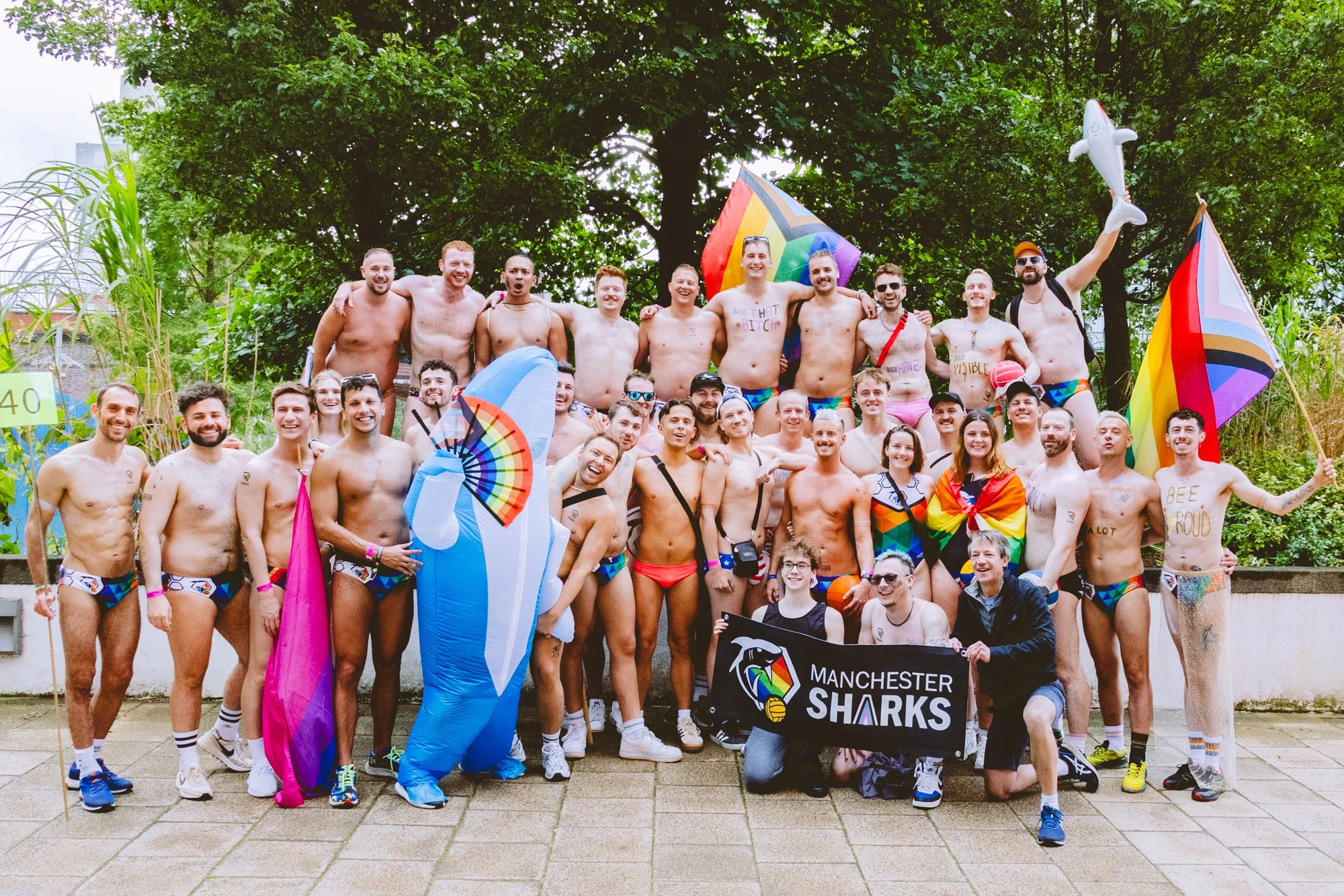 Group of shirtless men and women wearing colorful swimwear, celebrating at an outdoor pride event with rainbow flags, umbrellas, and a Manchester Shakers Sharks banner, standing in front of a large tree.