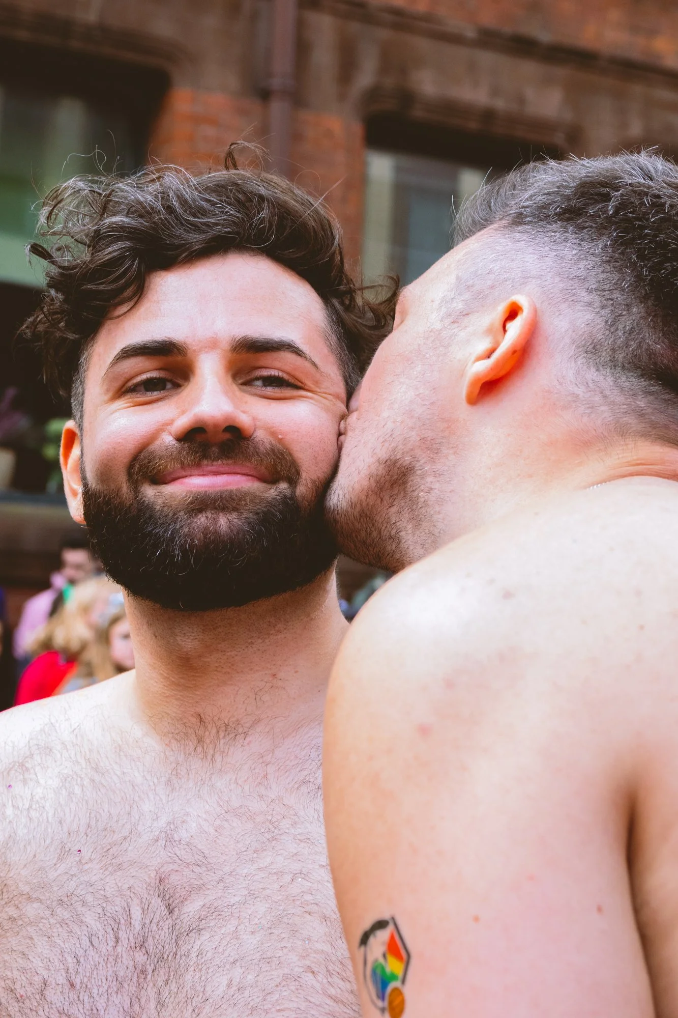 Two men in an outdoor setting with buildings in the background. One man with a beard and curly hair is smiling, while the other man is kissing his cheek. Both are shirtless, and the man on the right has a rainbow pride tattoo on his arm.