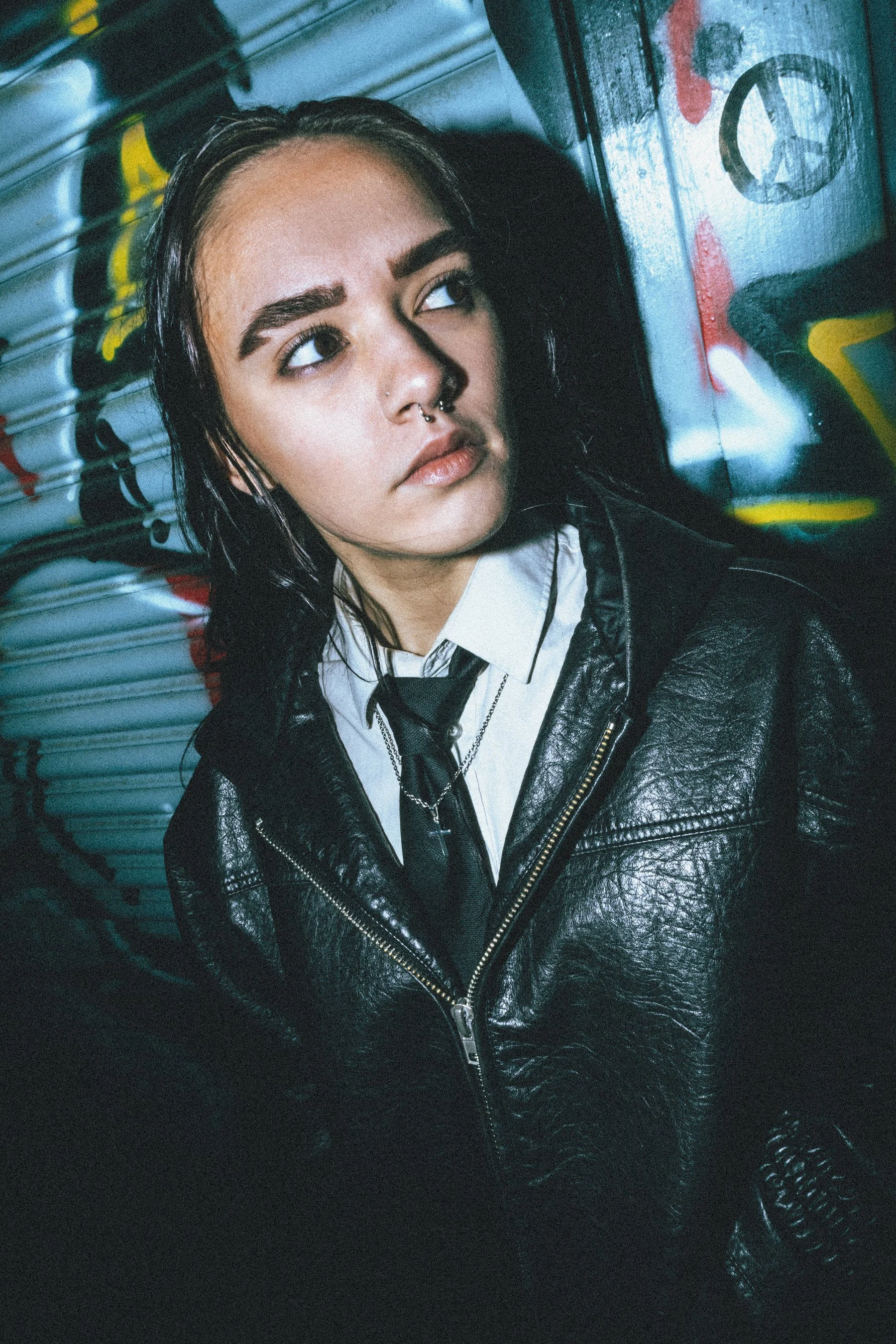 A young person with dark, wet hair and light makeup, including bold eyebrows and a septum piercing, leaning against a graffiti-covered metal wall at night. They wear a white shirt, a black tie, and a black leather jacket.