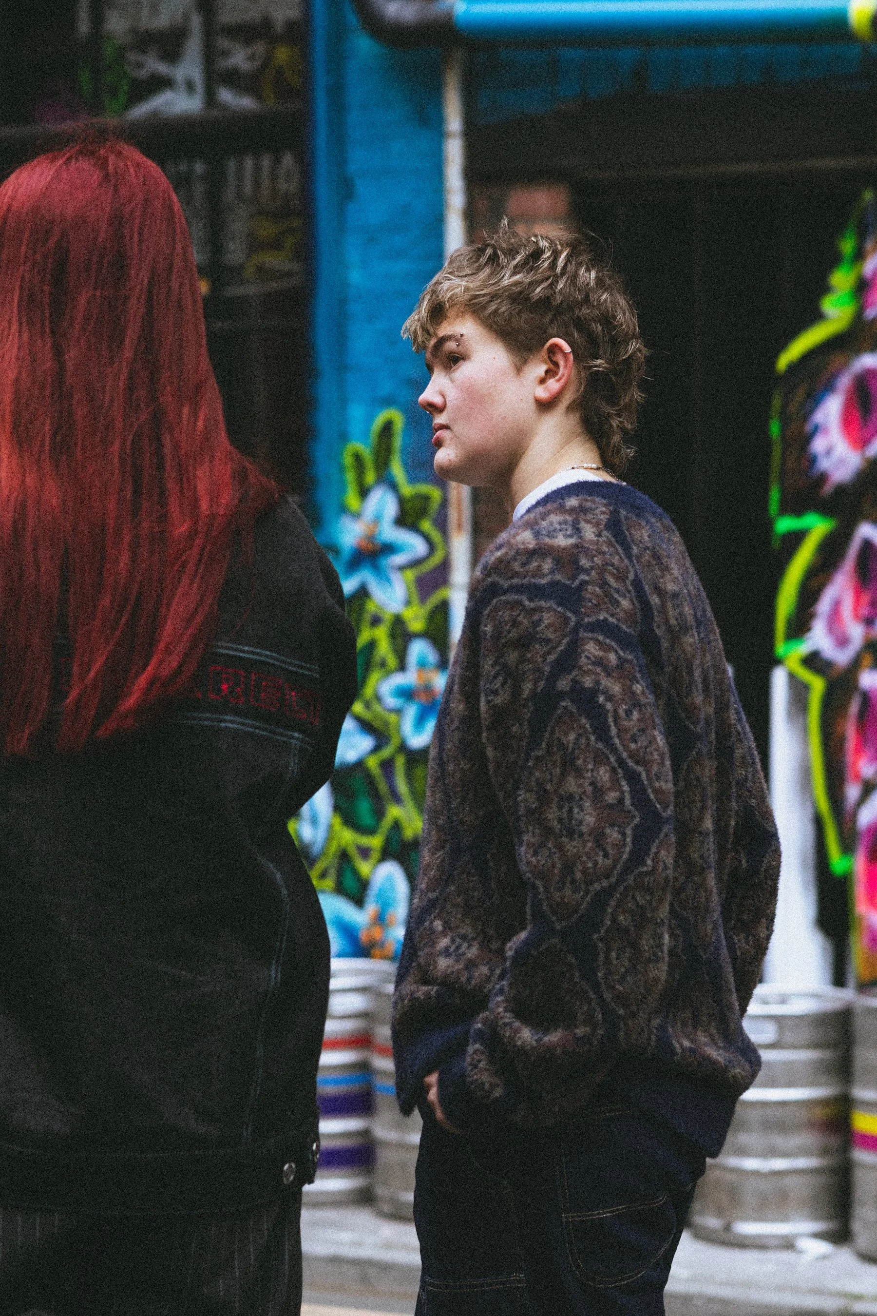 A person with short, wavy hair wearing a patterned jacket standing next to another person with long red hair.