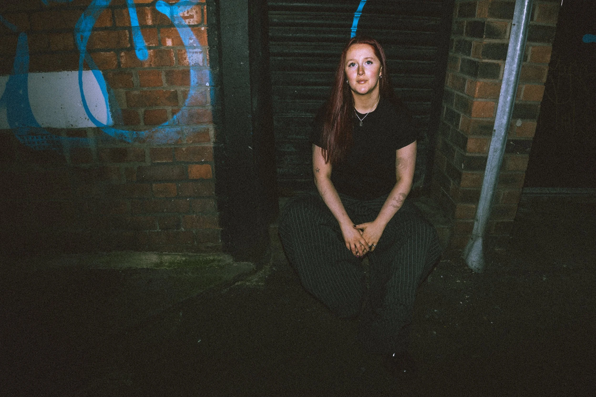 A young woman with red hair, wearing a black top and striped pants, sitting on the ground in an urban setting with brick walls and graffiti, looking at the camera.