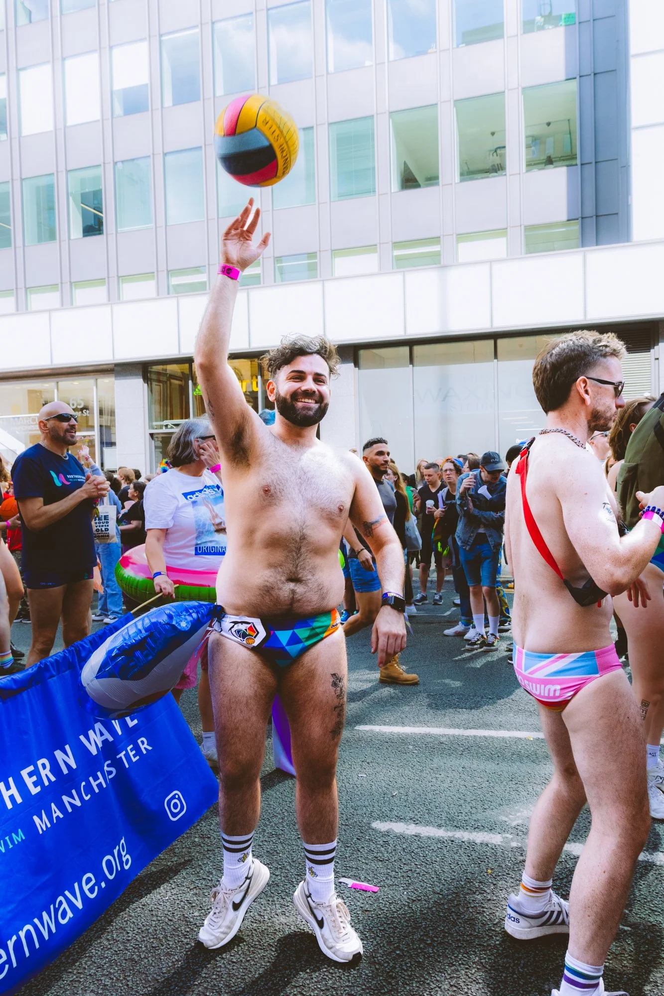 A man with a beard and rainbow-patterned shorts is smiling and holding a volleyball above his head during a parade or festival with a crowd of people around him, some in rainbow attire and accessories, in an urban setting.