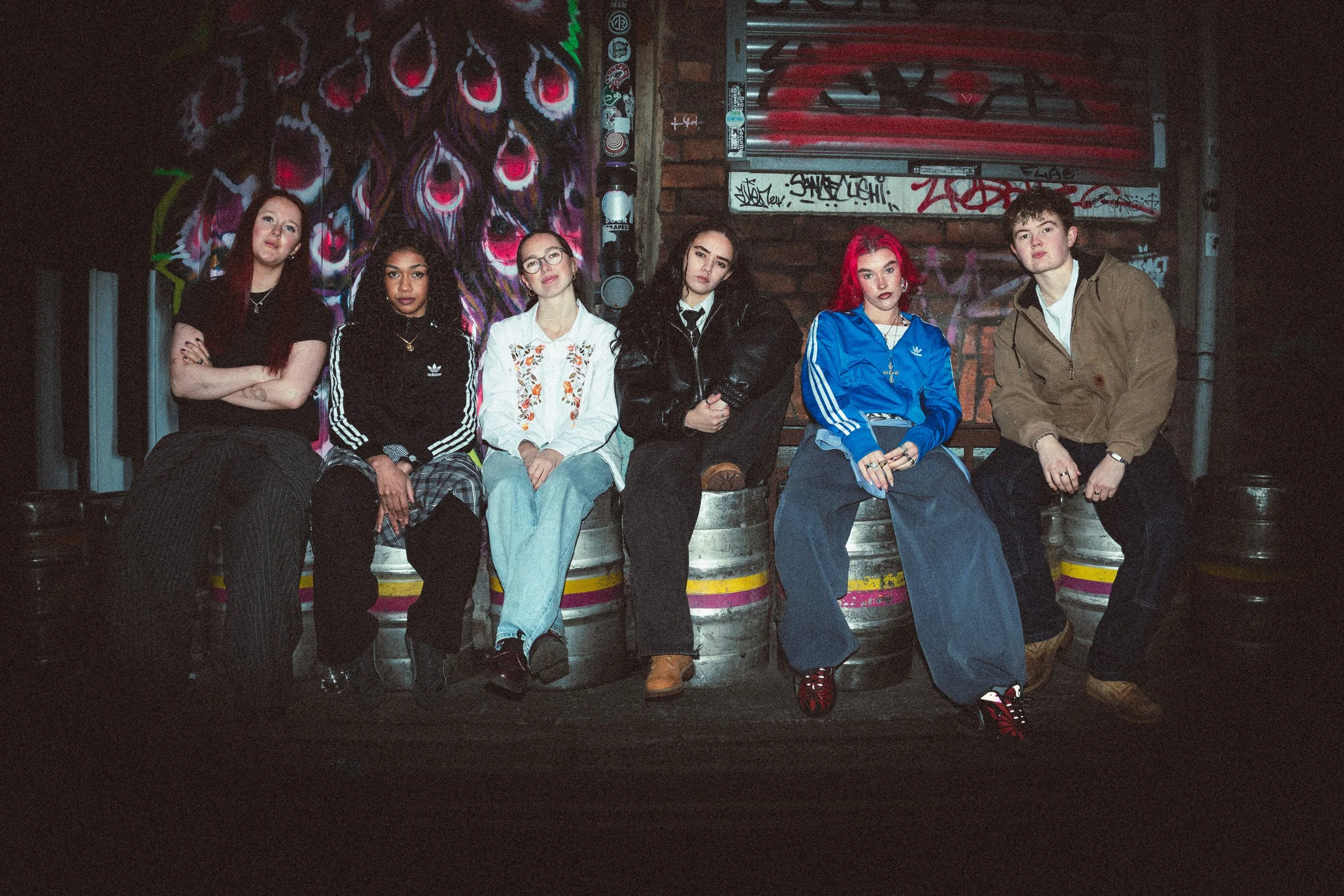 Six young people sitting on barrels with graffiti in an urban setting at night.