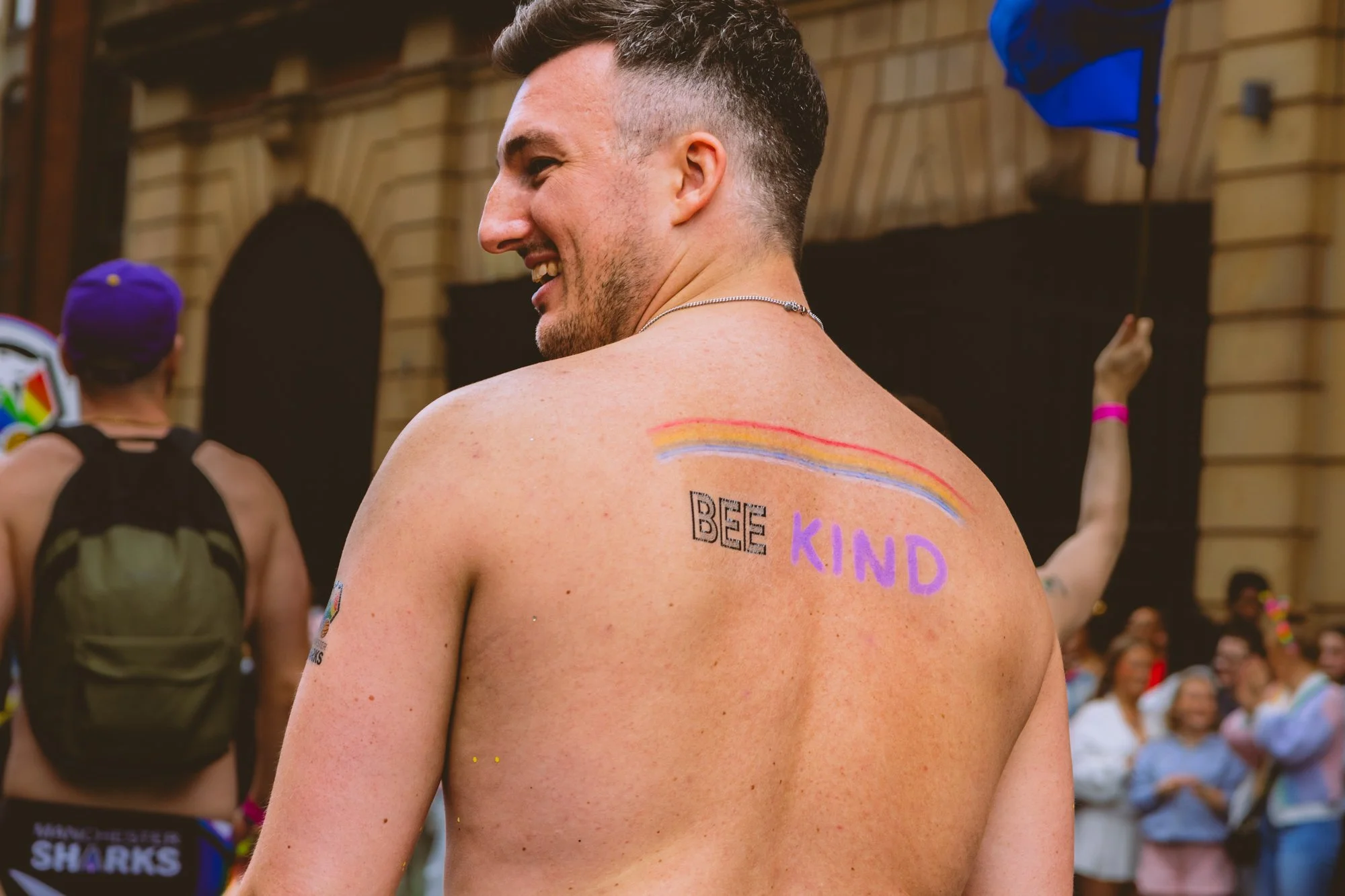 A smiling man with short dark hair and a beard, shirtless, at an event with a rainbow painted on his shoulder and the words "BE KIND" written on his back. There are people in the background, including a woman with a purple wristband and a rainbow fla