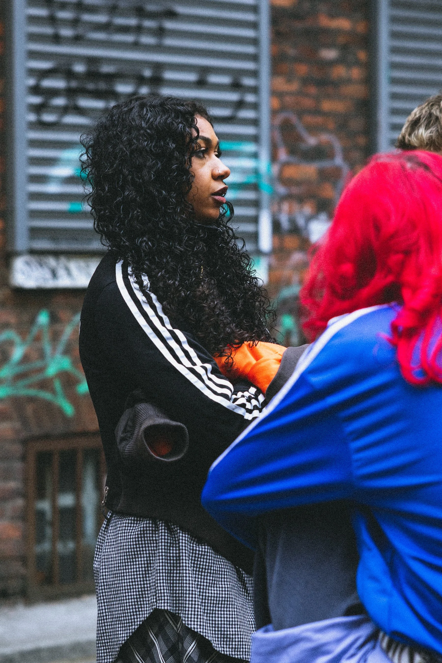 A person with long curly black hair, wearing a black jacket with white stripes on the sleeves, standing outdoors with a brick building and graffiti in the background. They are looking to the left.