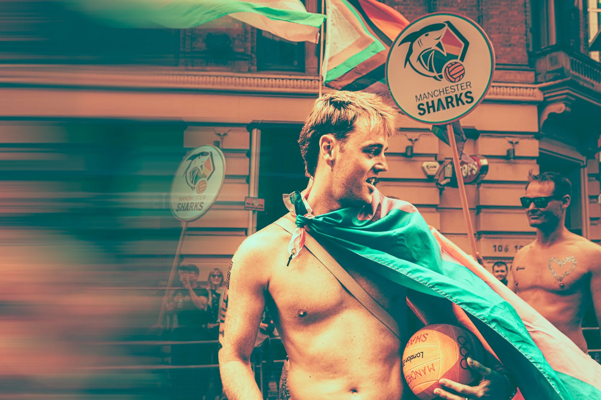 A shirtless man holding a volleyball with the Manchester Sharks logo during a parade or celebration on a city street. He is draped in a flag, and other people are seen in the background, some wearing sunglasses. There are signs and buildings visible.