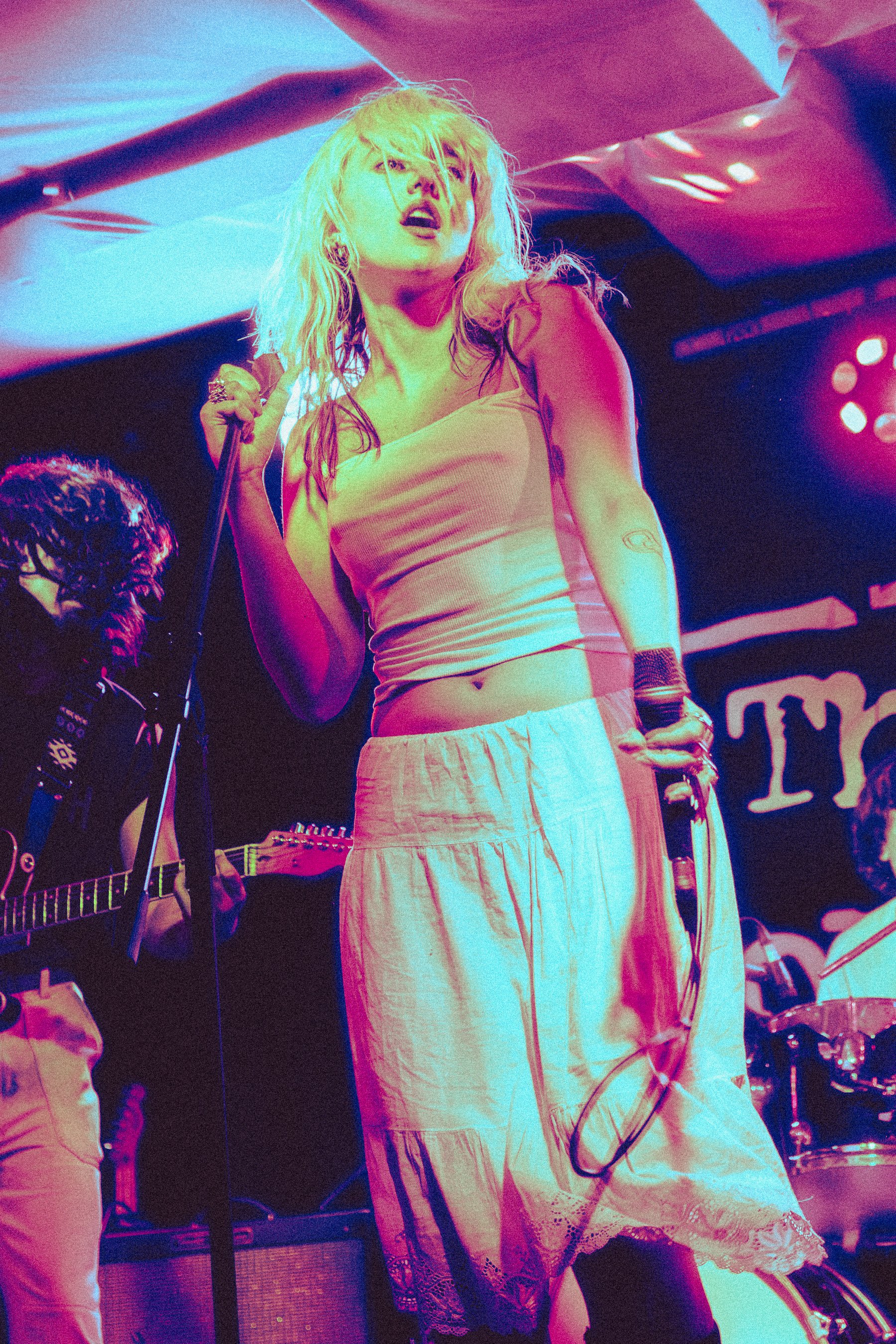 A woman with blonde hair singing or performing on stage, holding a microphone, with colorful stage lights and musical instruments in the background.
