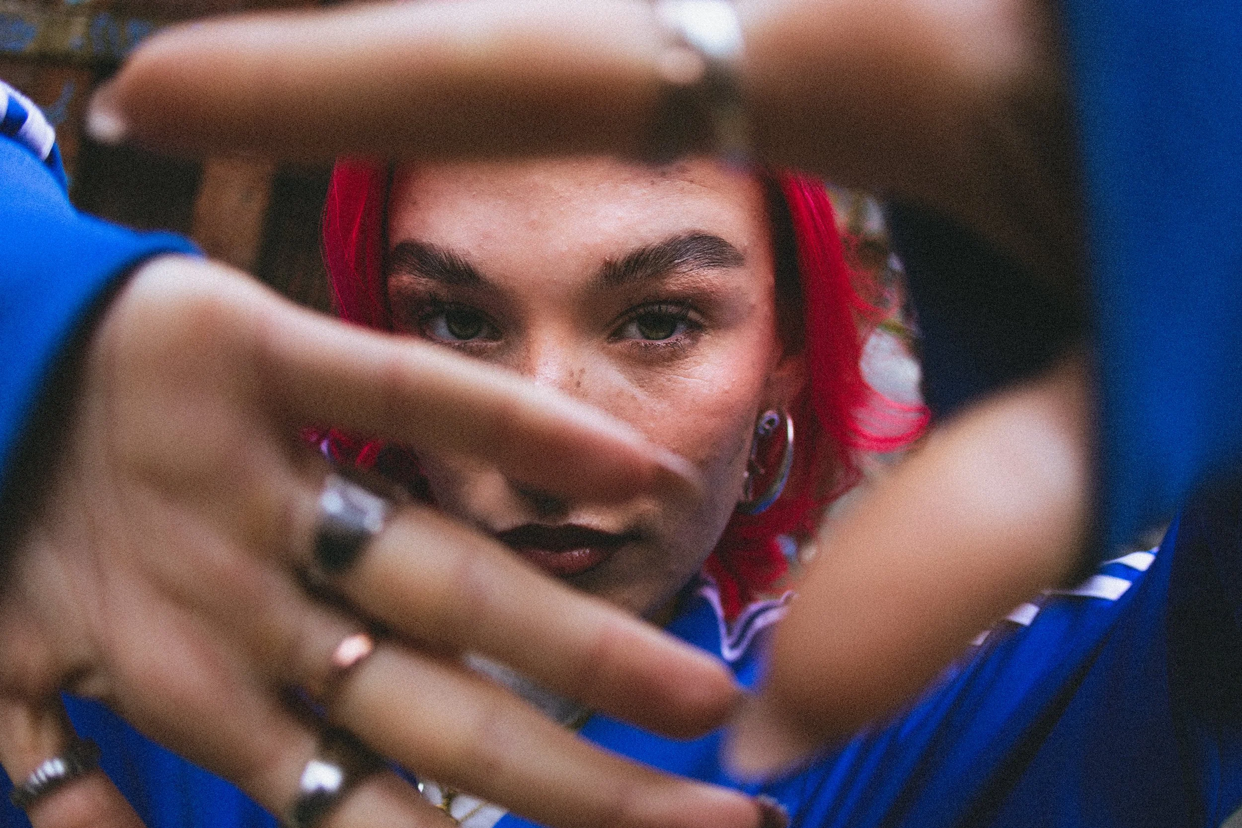 A woman with red hair and fair skin, wearing makeup with dark lipstick, is looking at the camera through her fingers, which are decorated with rings, with a playful or mischievous expression.