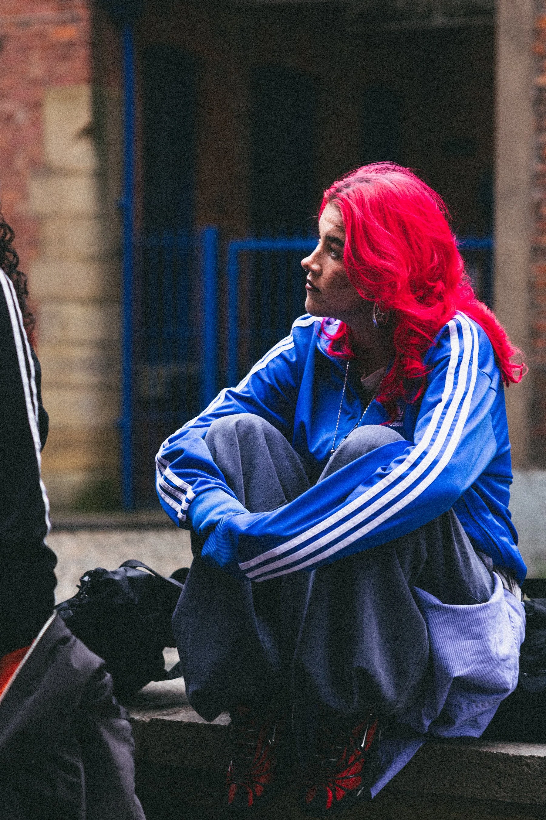 A person with bright red hair sitting on a curb and talking to a person off-frame, wearing a blue Adidas jacket with white stripes, grey sweatpants, and red and black sneakers.