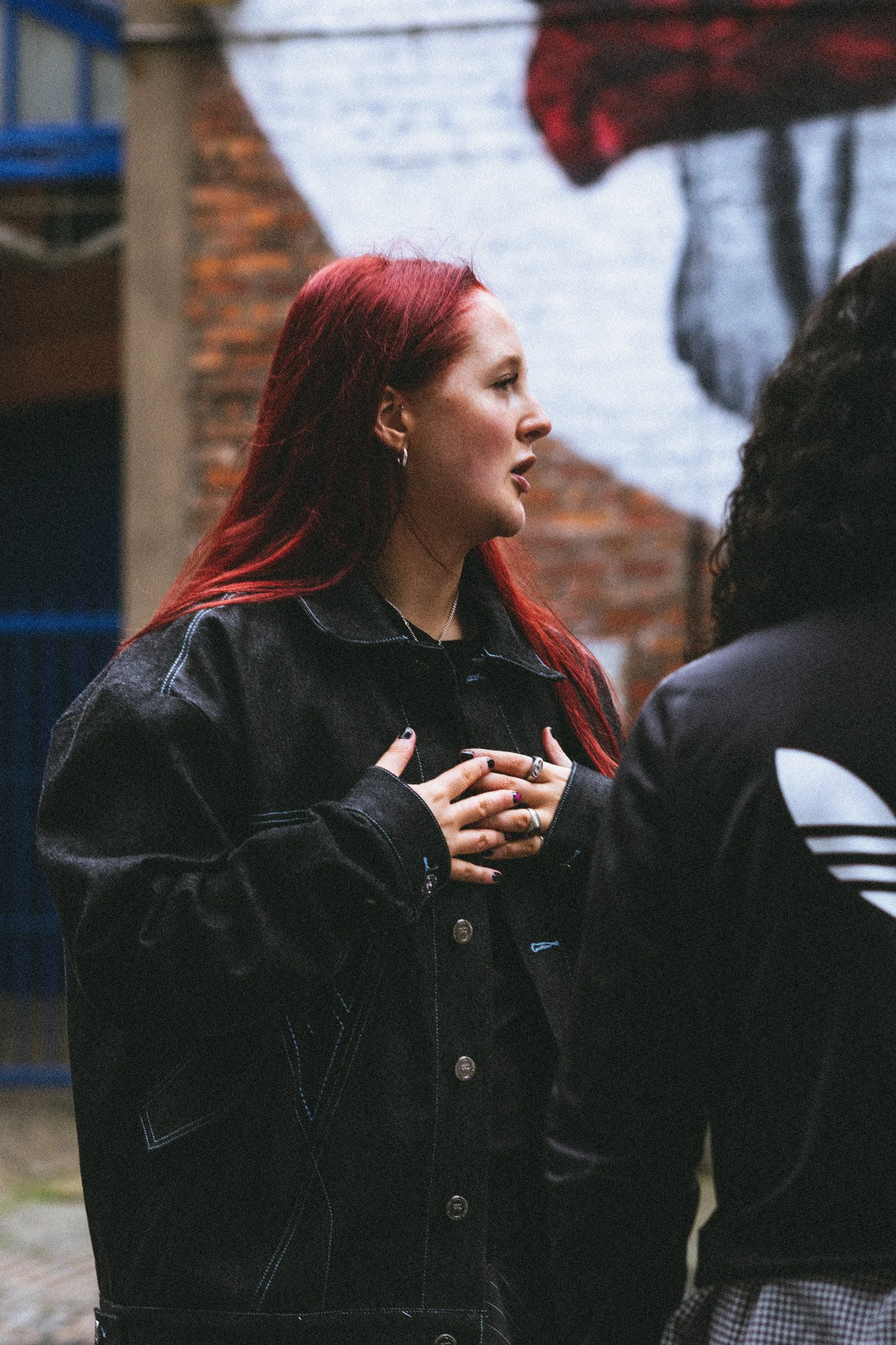 A person with long red hair, wearing a black denim jacket, is talking to someone outside the frame. They have their hands on her chest and appears focused on the conversation. 